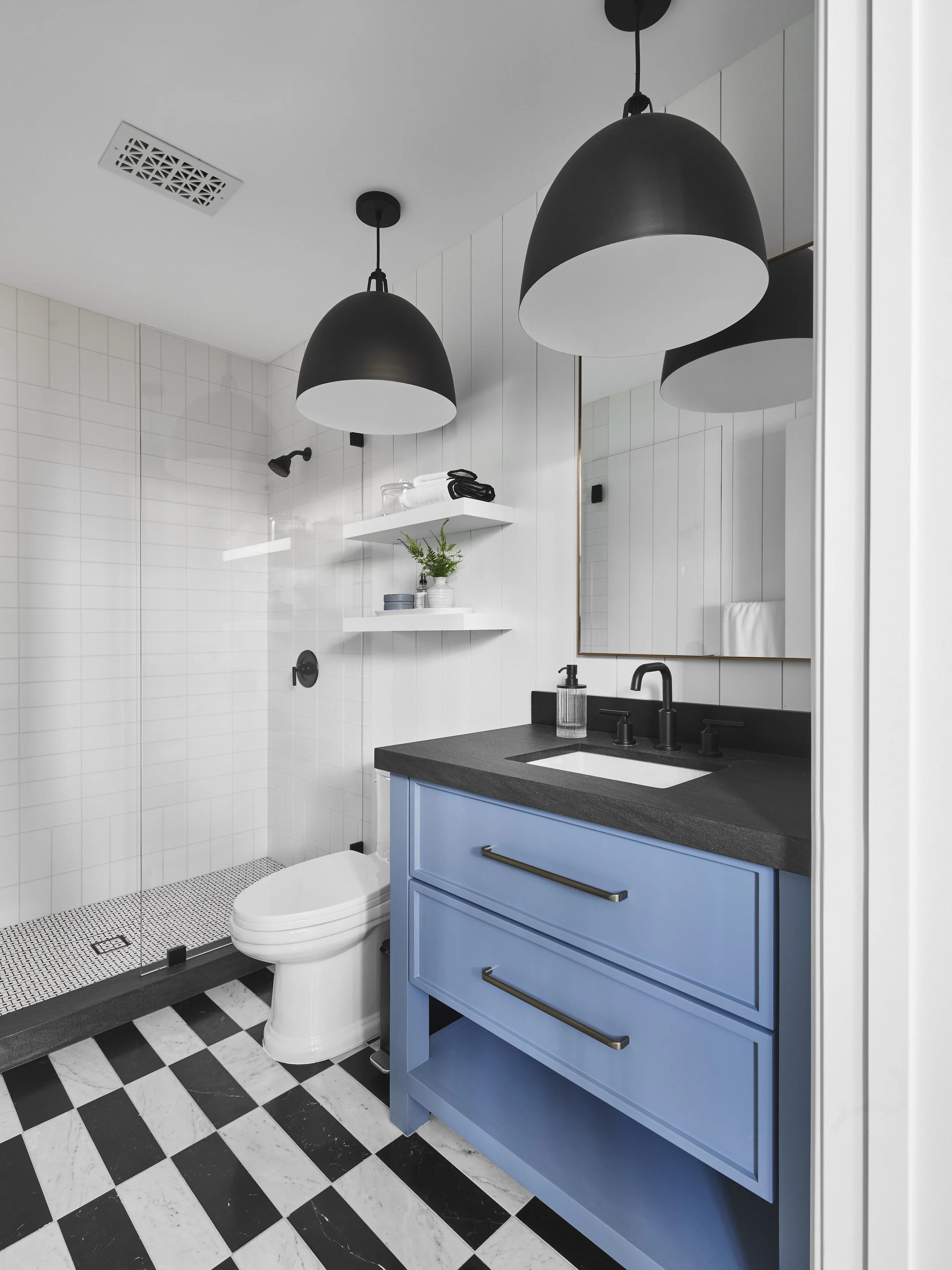 Modern bathroom with black and white checkered floor, blue vanity with black countertop, black faucet, and hanging black light fixtures. Features a walk-in shower with glass door, white tiled walls, and white shelving with decor items.