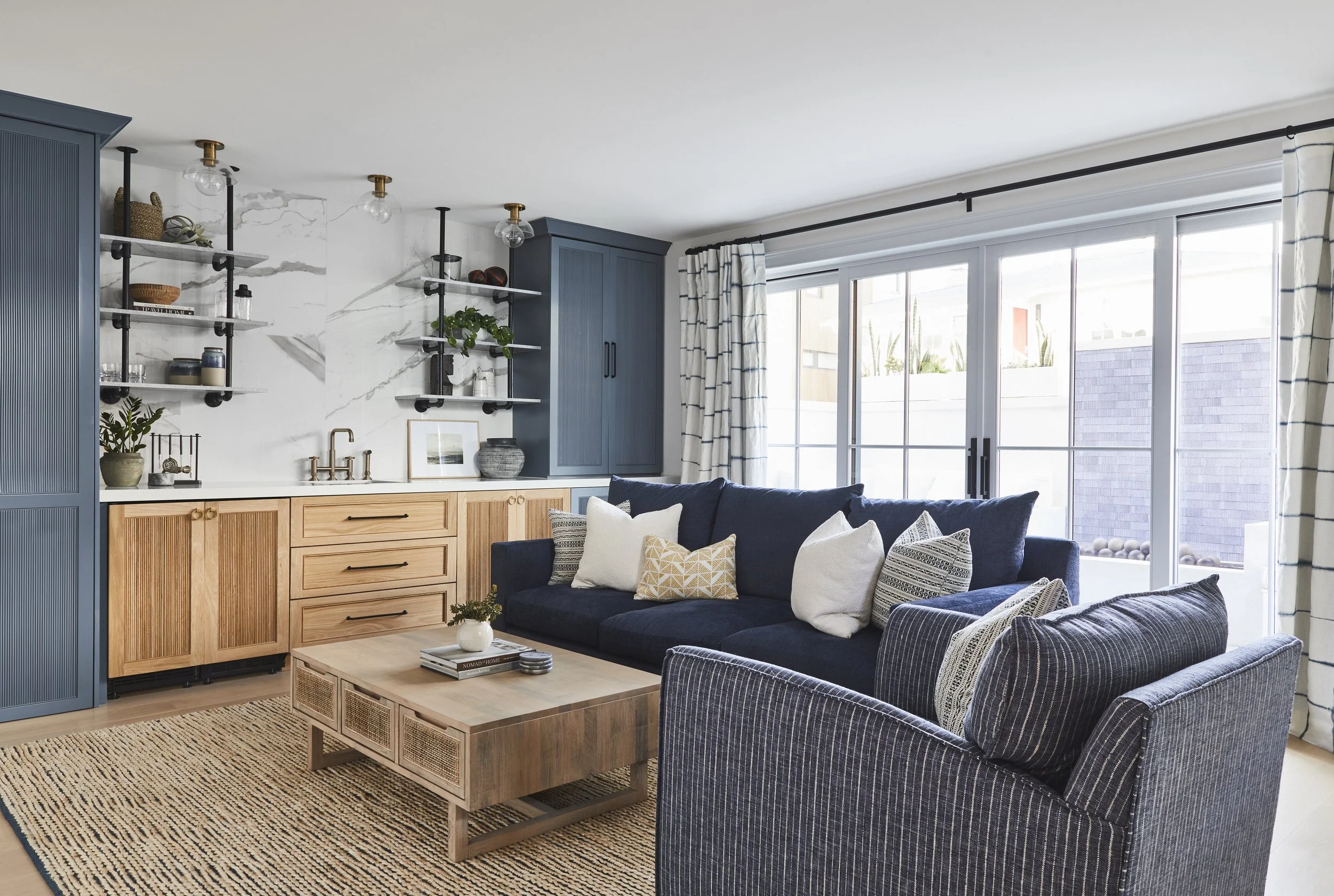 Modern living room with a navy blue sofa, decorative pillows, wooden coffee table, built-in cabinets, shelves with decor items, and large windows with striped curtains.