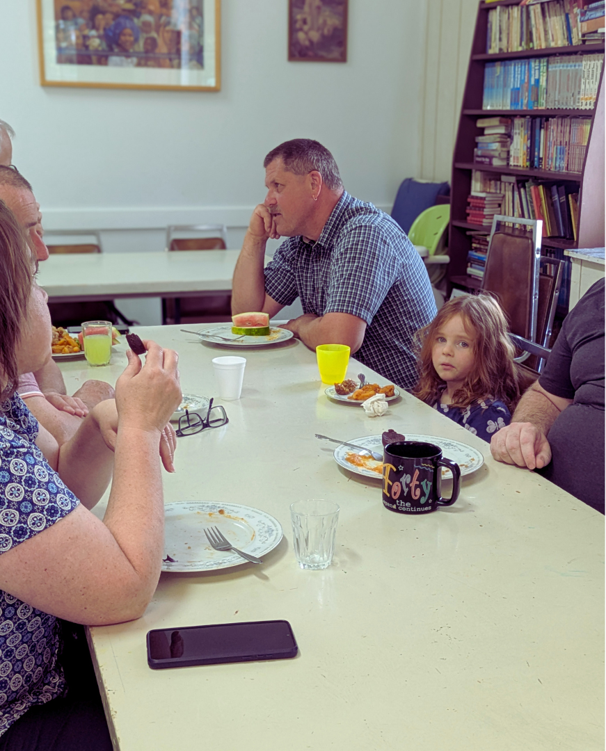 A group of people sitting around a beige dining table with plates of food, one person eating, and a young girl sitting next to a man. The room has bookshelves and framed pictures on the wall.