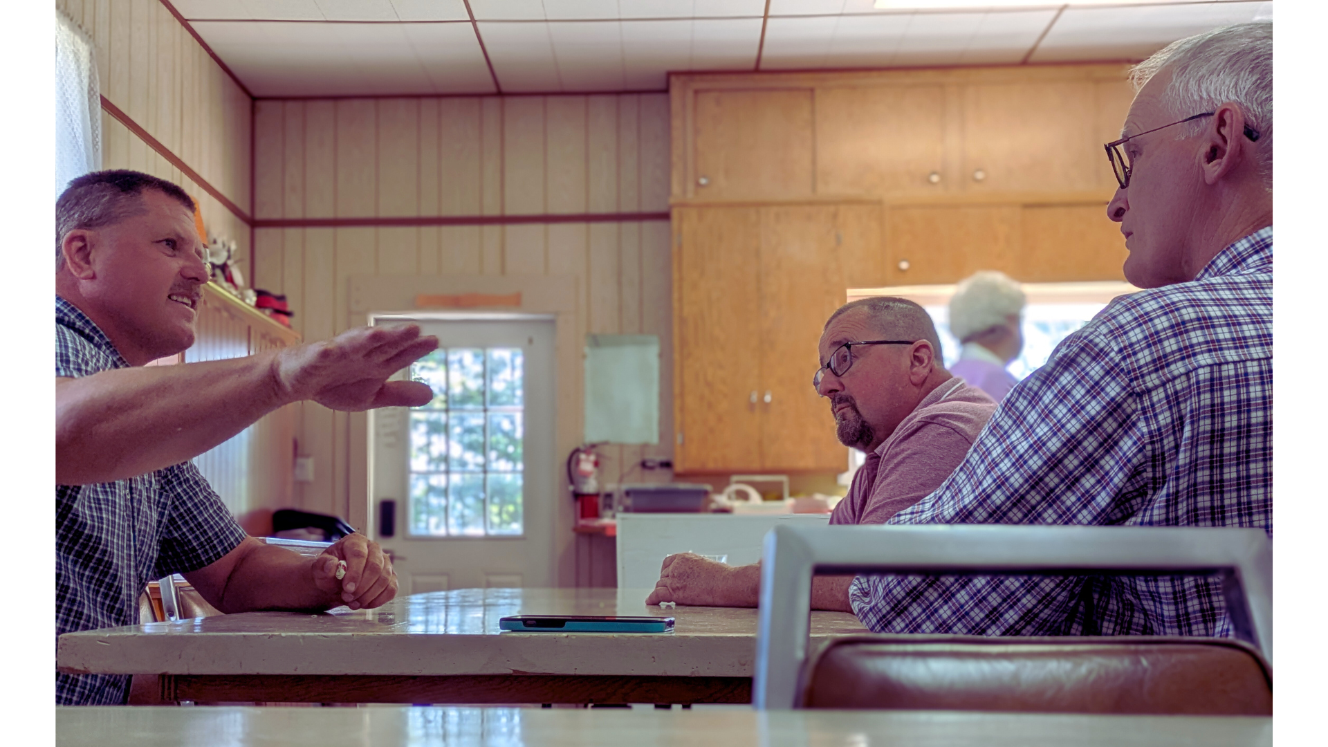 Three men sitting at a table in a kitchen, engaged in conversation. The man on the left is gesturing with his hand and smiling. The man in the middle has glasses and a beard, and the man on the right has white hair and glasses.