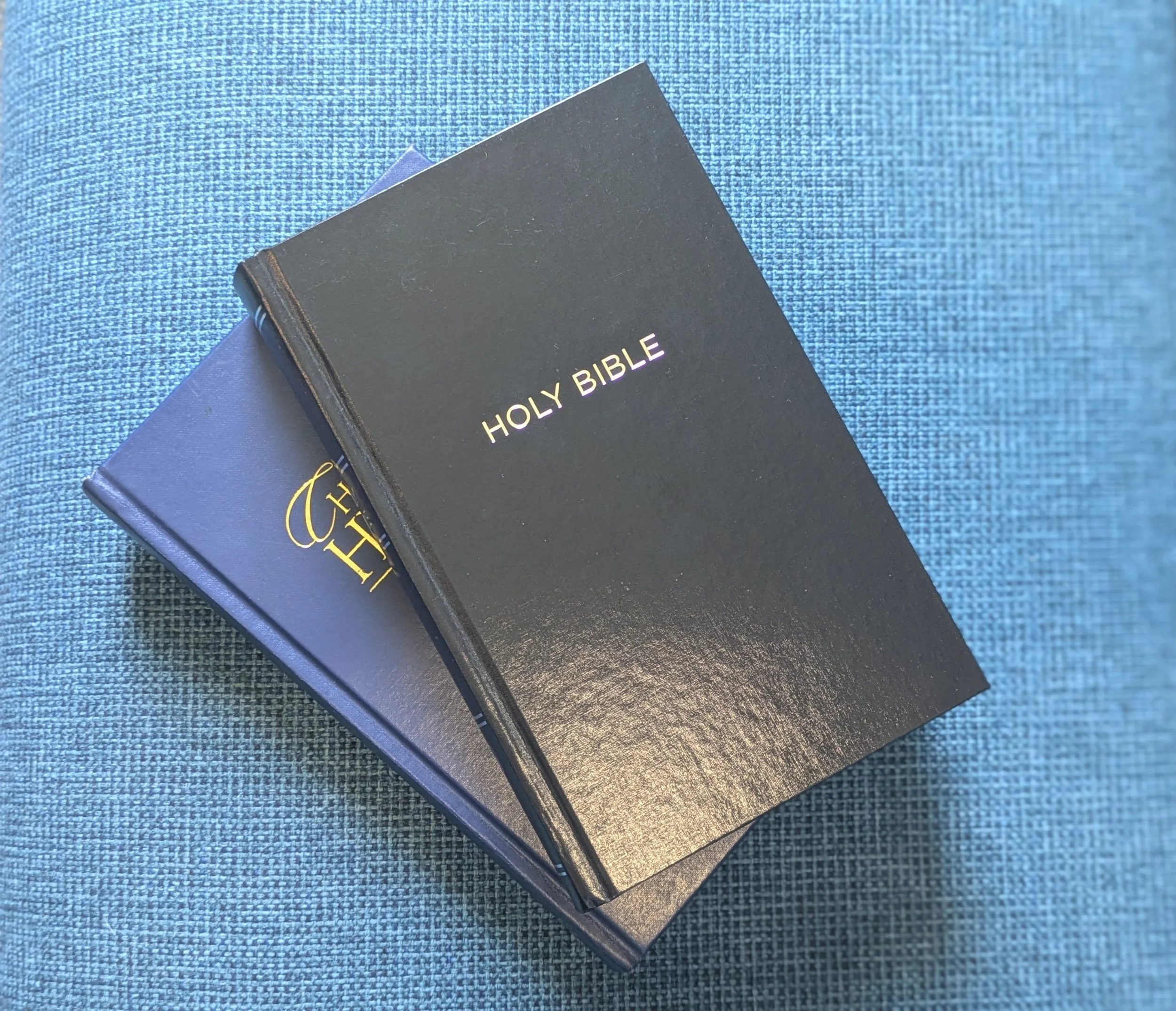 Two Bibles, one labeled 'HOLY BIBLE' on top, resting on a blue textured fabric surface.