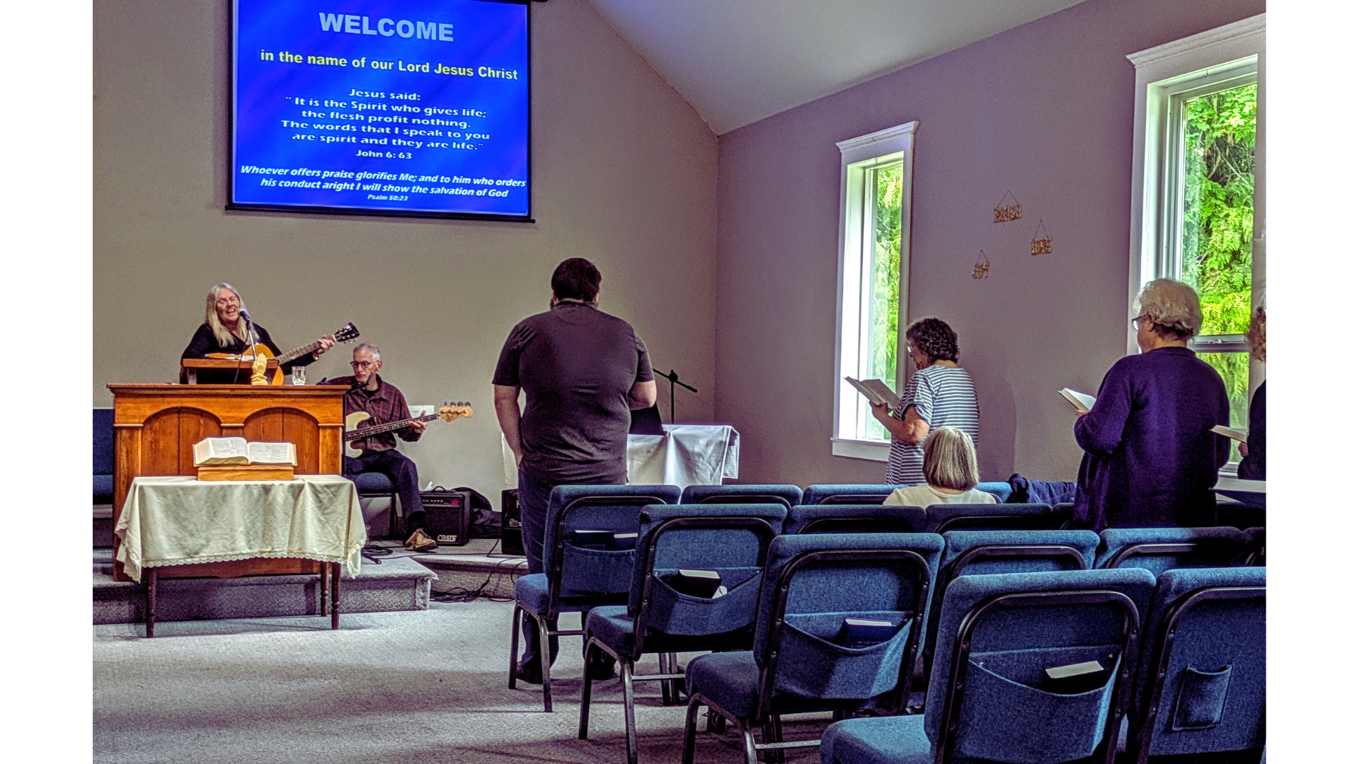 A group of people in a church or worship space, standing and singing, with a woman playing guitar and a man playing bass guitar at the front, facing a large screen displaying religious text and lyrics.