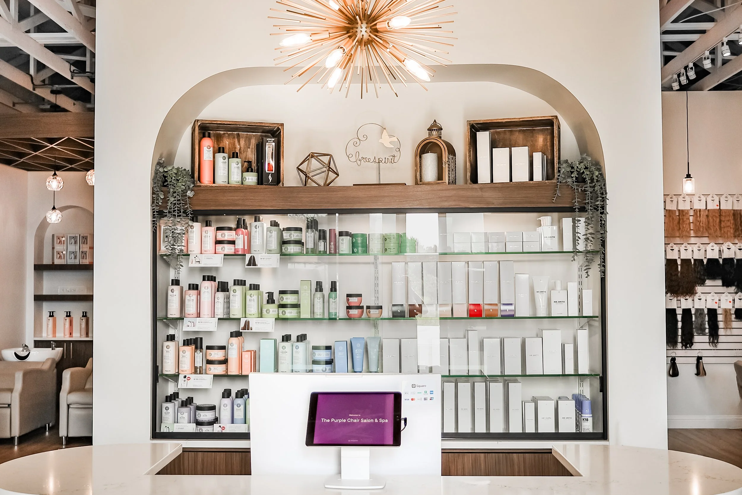 Display of skincare or beauty products on shelves in a salon or spa, with decorative items and a chandelier above, and a reception desk with a purple screen in the foreground.