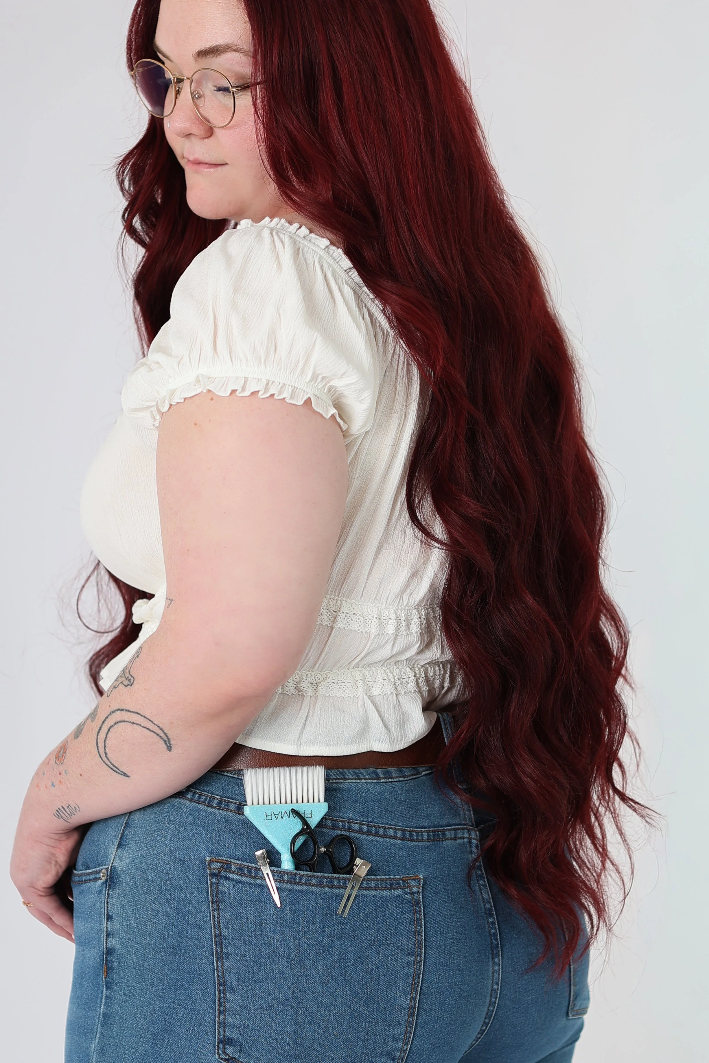 Journey Bryant with long, wavy red hair wearing glasses and a white blouse, standing with her back partially facing the camera, with tattoos on her left arm, holding hairdressing scissors and related tools in her back pocket.