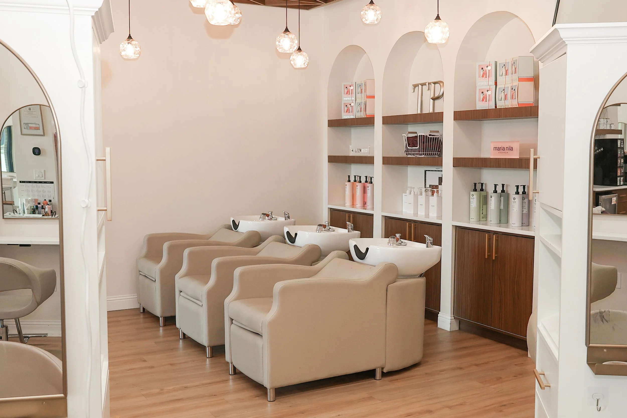 A row of beige salon chairs with white sinks in front of them, set against a wall with shelves containing hair care products and decorative items in a salon.