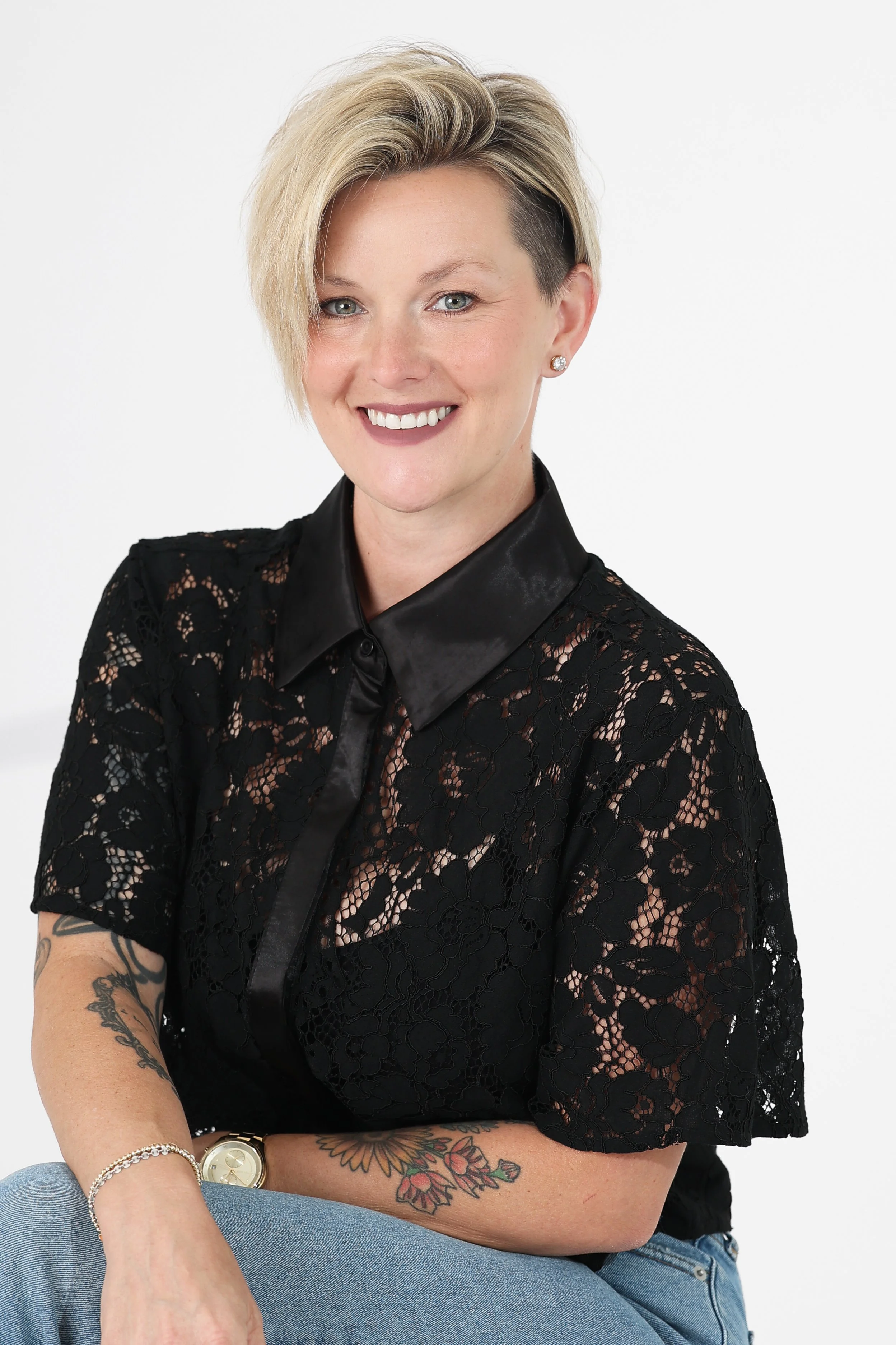 Brittany Wilson with short blonde hair and tattoos on her arm, wearing a black lace top and a wristwatch, sitting with crossed arms and smiling against a white background.