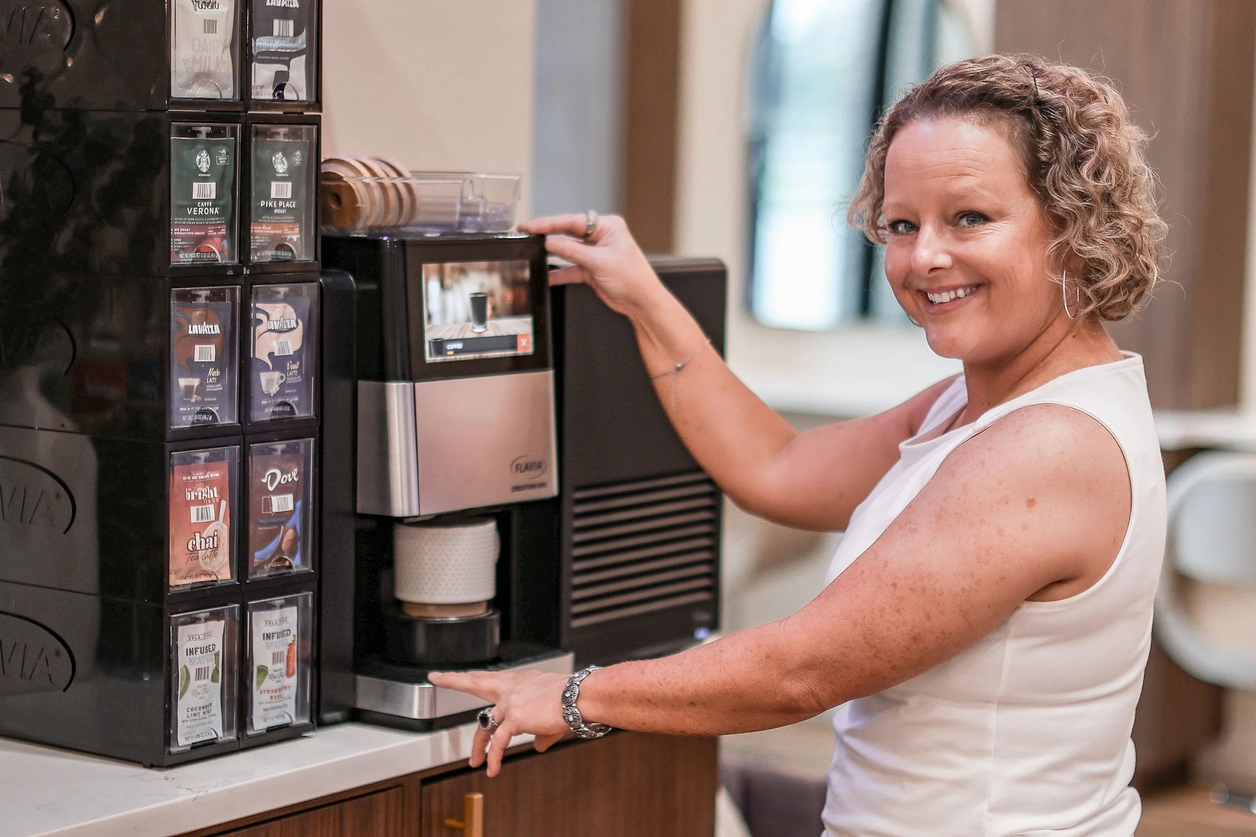 A woman with curly blonde hair smiling as she operates a coffee and tea vending machine in a room with natural light.