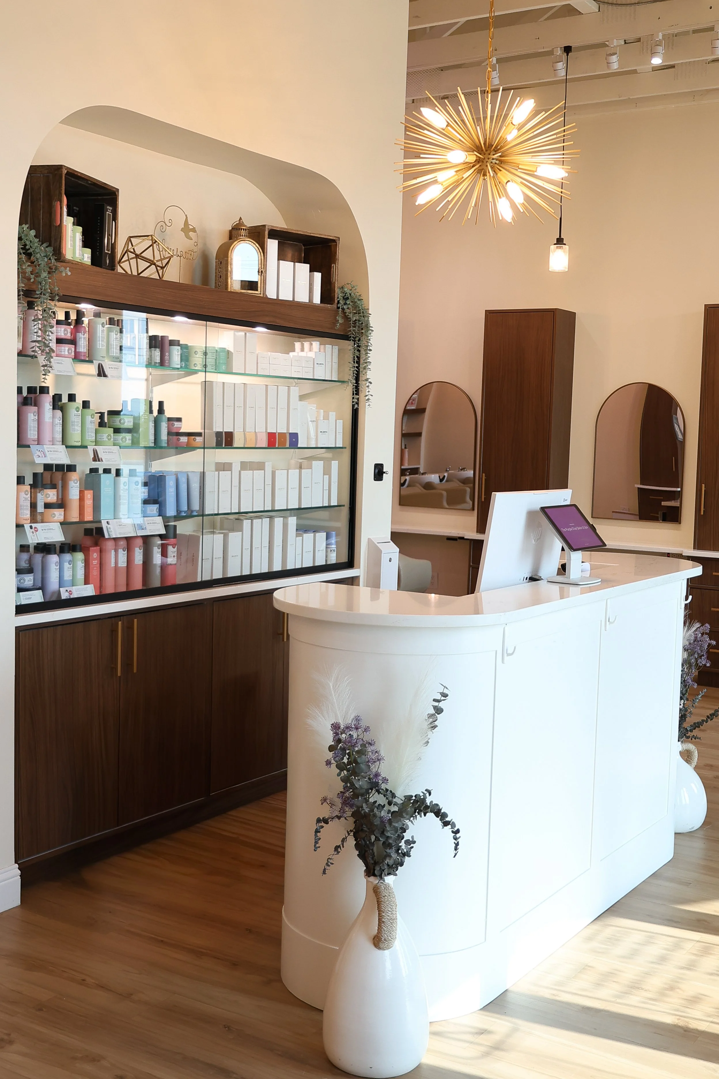 Interior of a modern retail store with a white curved checkout counter, wooden flooring, and shelves displaying skincare or beauty products. The background features mirrors, wooden cabinets, and decorative lighting fixtures, including a starburst cha