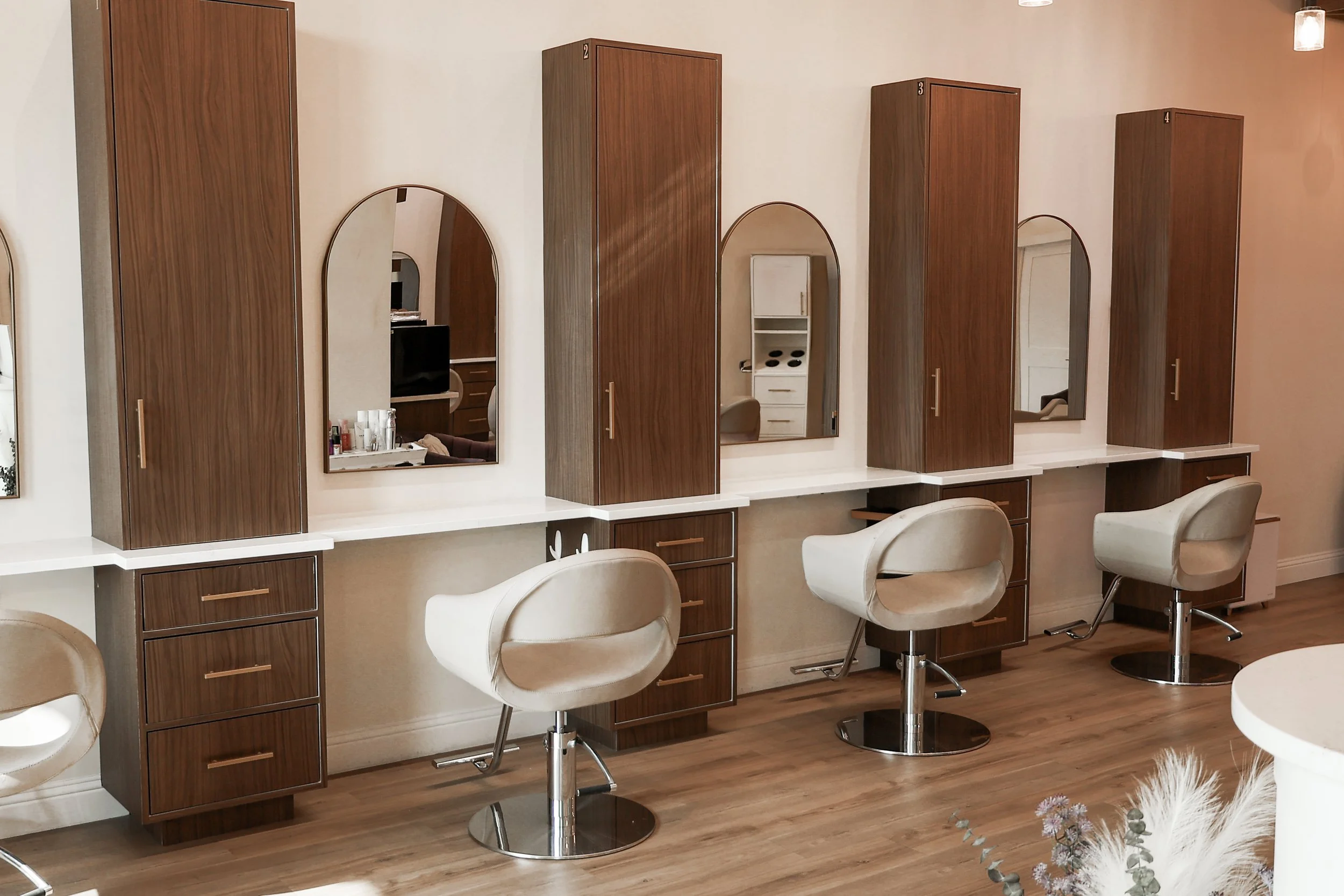 A row of white salon chairs with brown wooden cabinets and mirrors behind them, set against a beige wall with hardwood flooring.