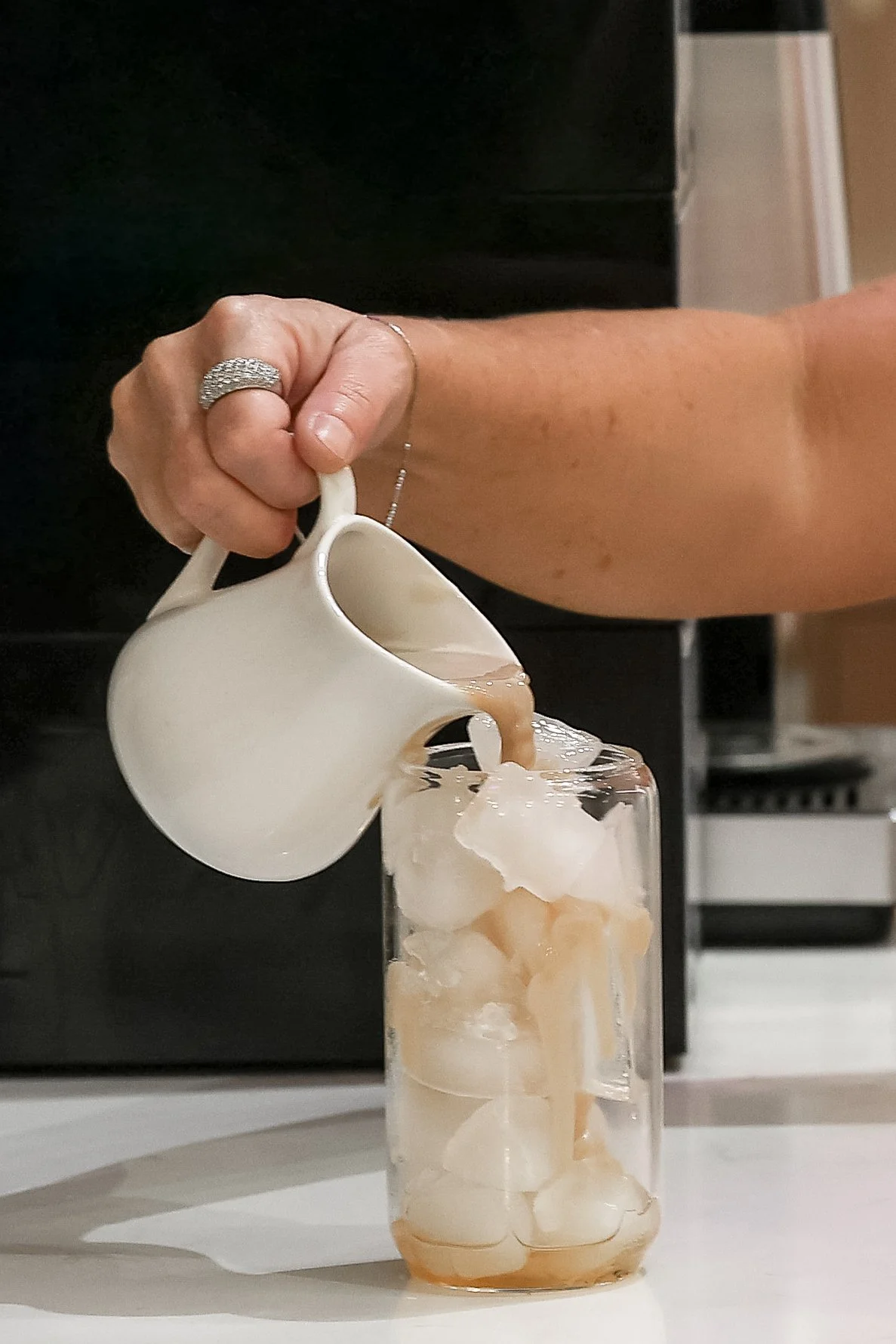 A person is pouring coffee from a white cream pitcher into a tall glass filled with ice cubes.