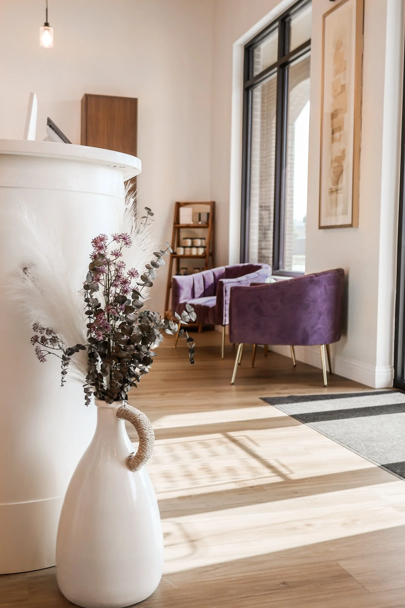 Interior of a modern room with a white vase filled with dried flowers and eucalyptus in the foreground, purple armchairs near large window, a small wooden ladder shelf with candles, a wooden cabinet, and framed artwork on the wall, natural sunlight s