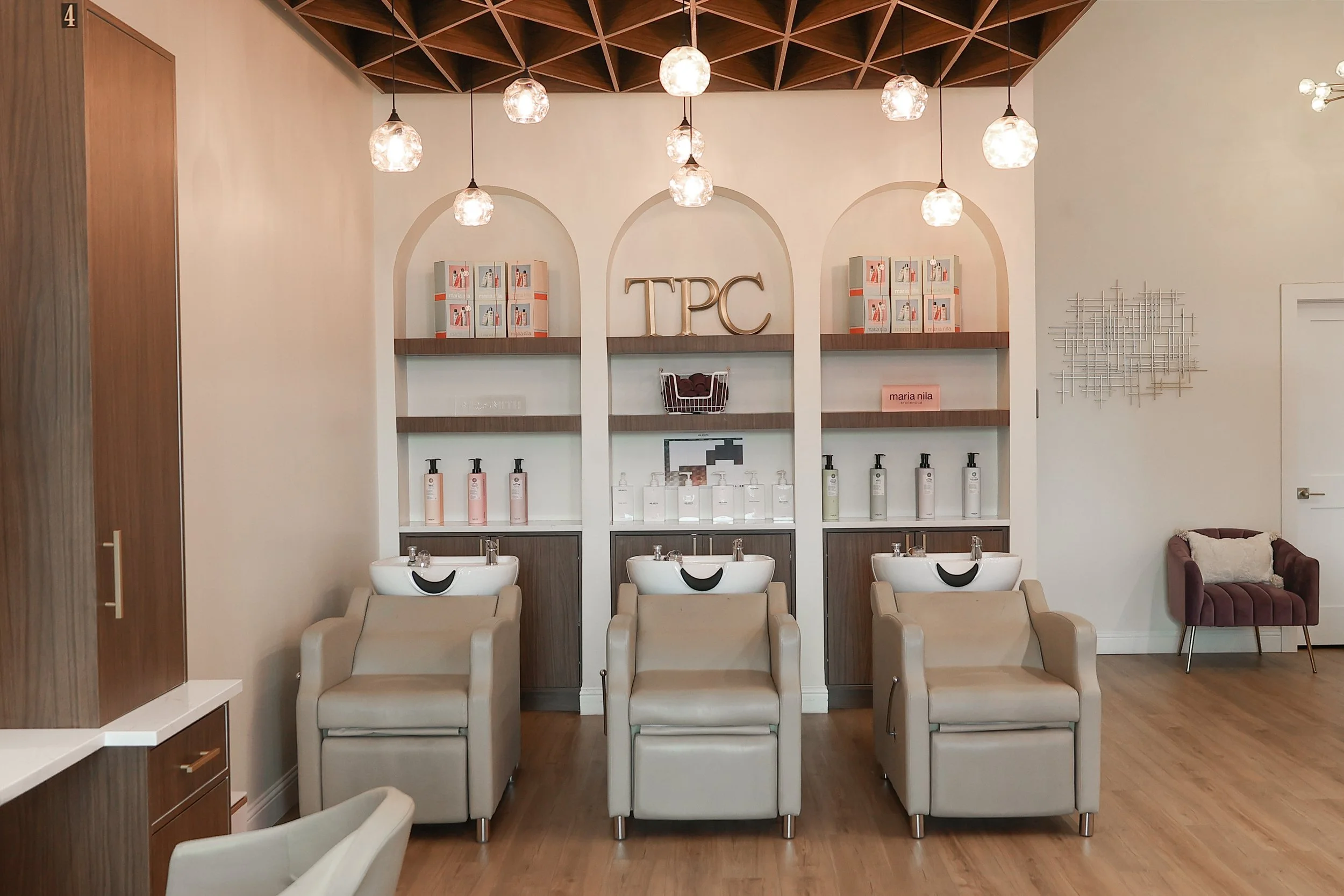 A modern salon or spa room with three white sinks and chairs, shelves holding hair and skincare products, decorative wooden wall art, illuminated hanging lights, and styled wood ceiling.