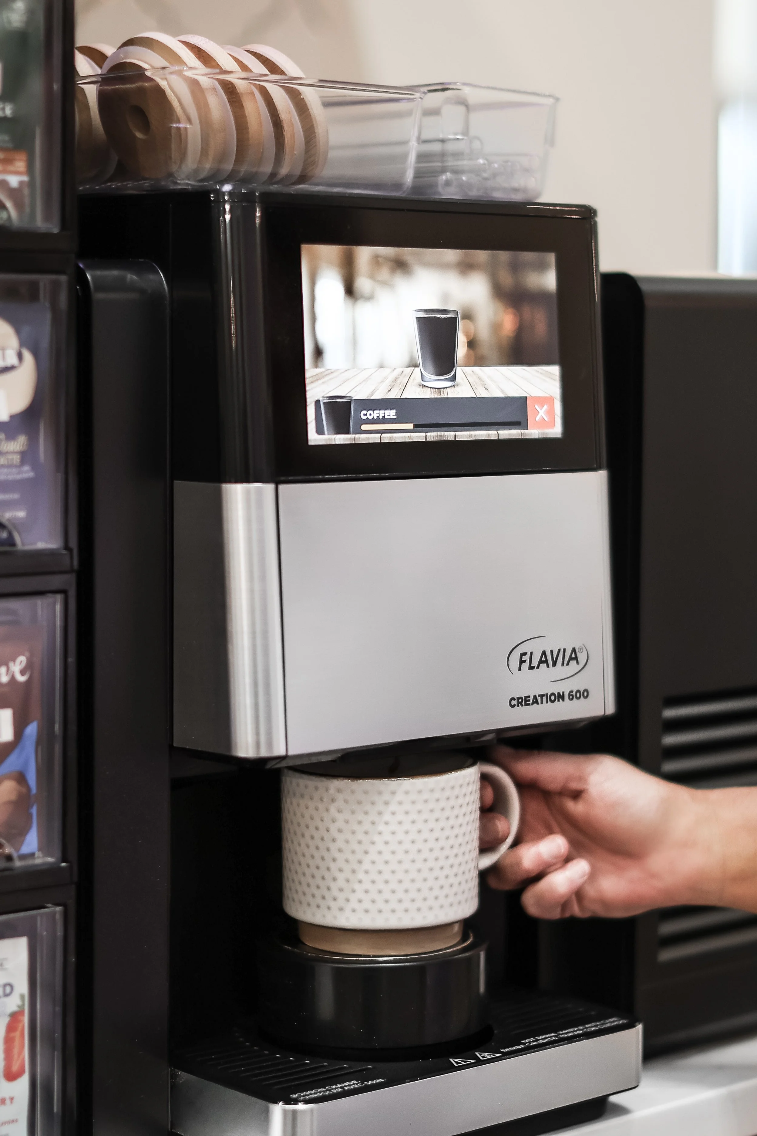 A person inserting a mug into a Flavia Creation 600 coffee machine with a digital screen showing a coffee icon.