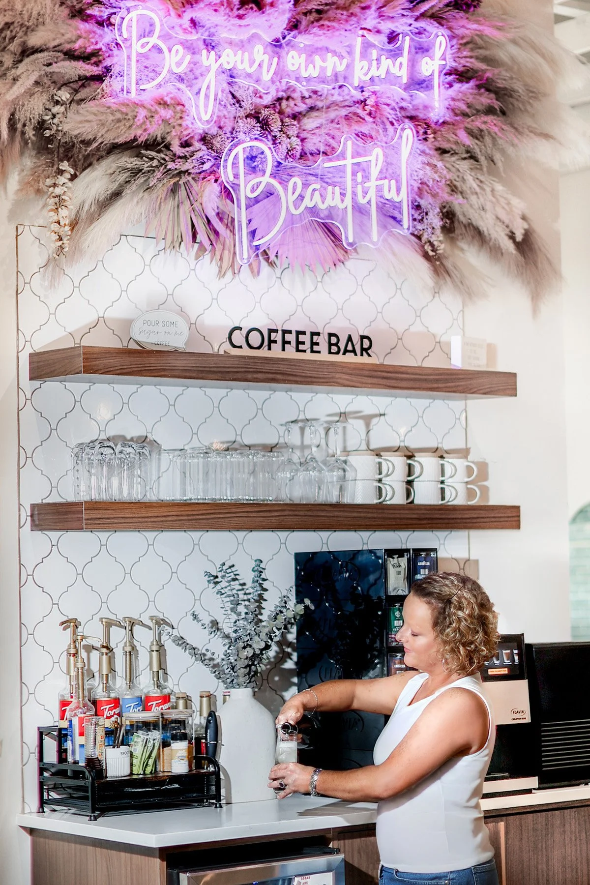A woman with curly hair wearing a white sleeveless top is working behind a coffee bar, filling a glass. The coffee bar has a white countertop and a decorative backsplash with a white and gray geometric tile pattern. There are shelves above the counte