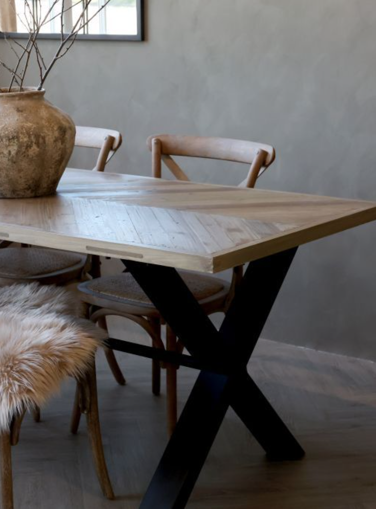 Mesa de madera con patas negras y una gran maceta con ramas secas en el centro, acompañada de sillas de madera y una de ellas con asiento de piel de animal.