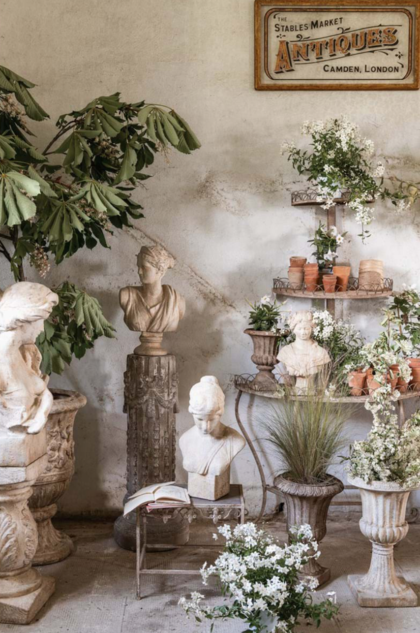 Estantería con macetas de barro, plantas y esculturas clásicas de piedra en un ambiente de mercado de antigüedades vintage.