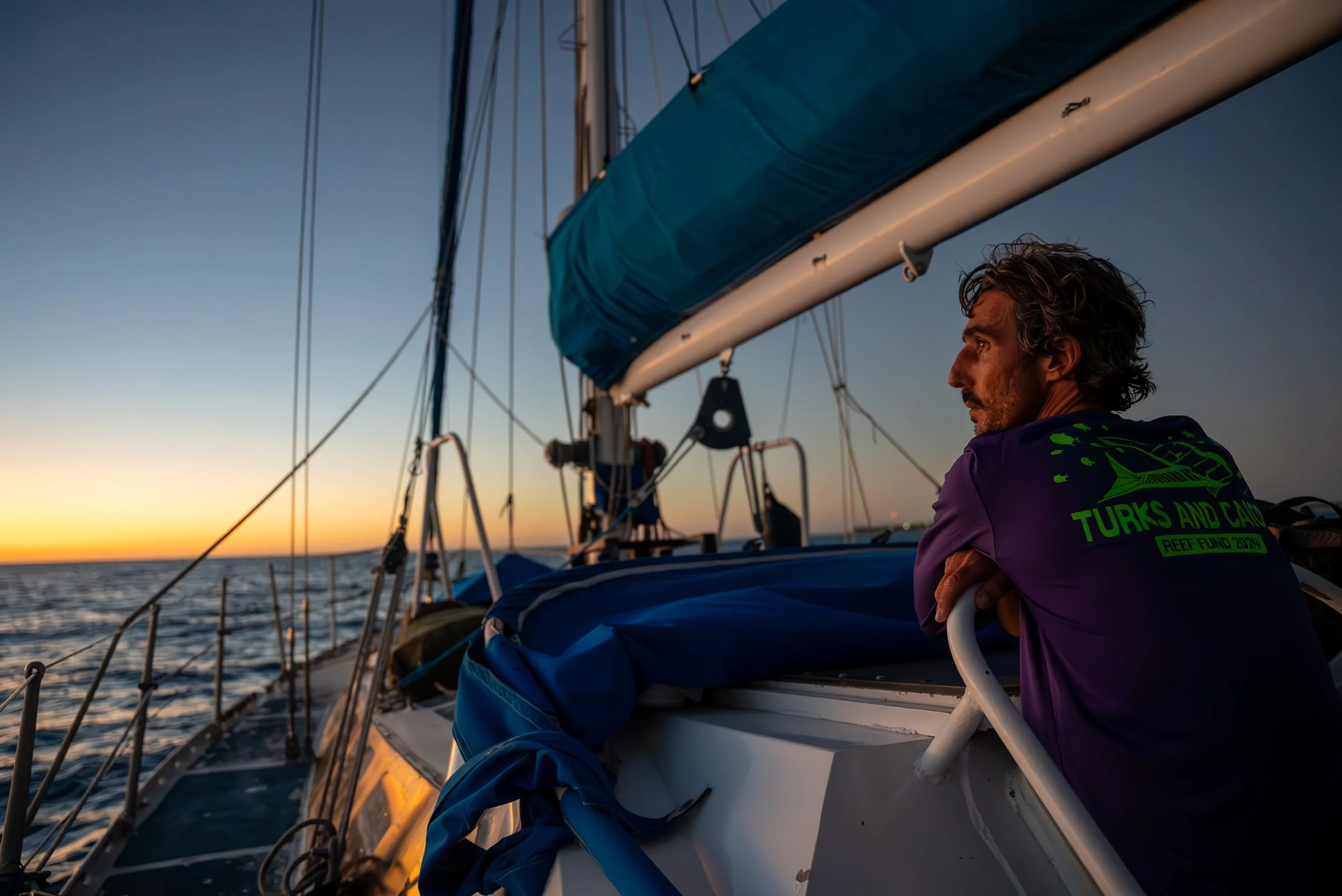 A man with gray hair and a beard sitting on a sailboat during sunset, looking out at the horizon over calm ocean water.