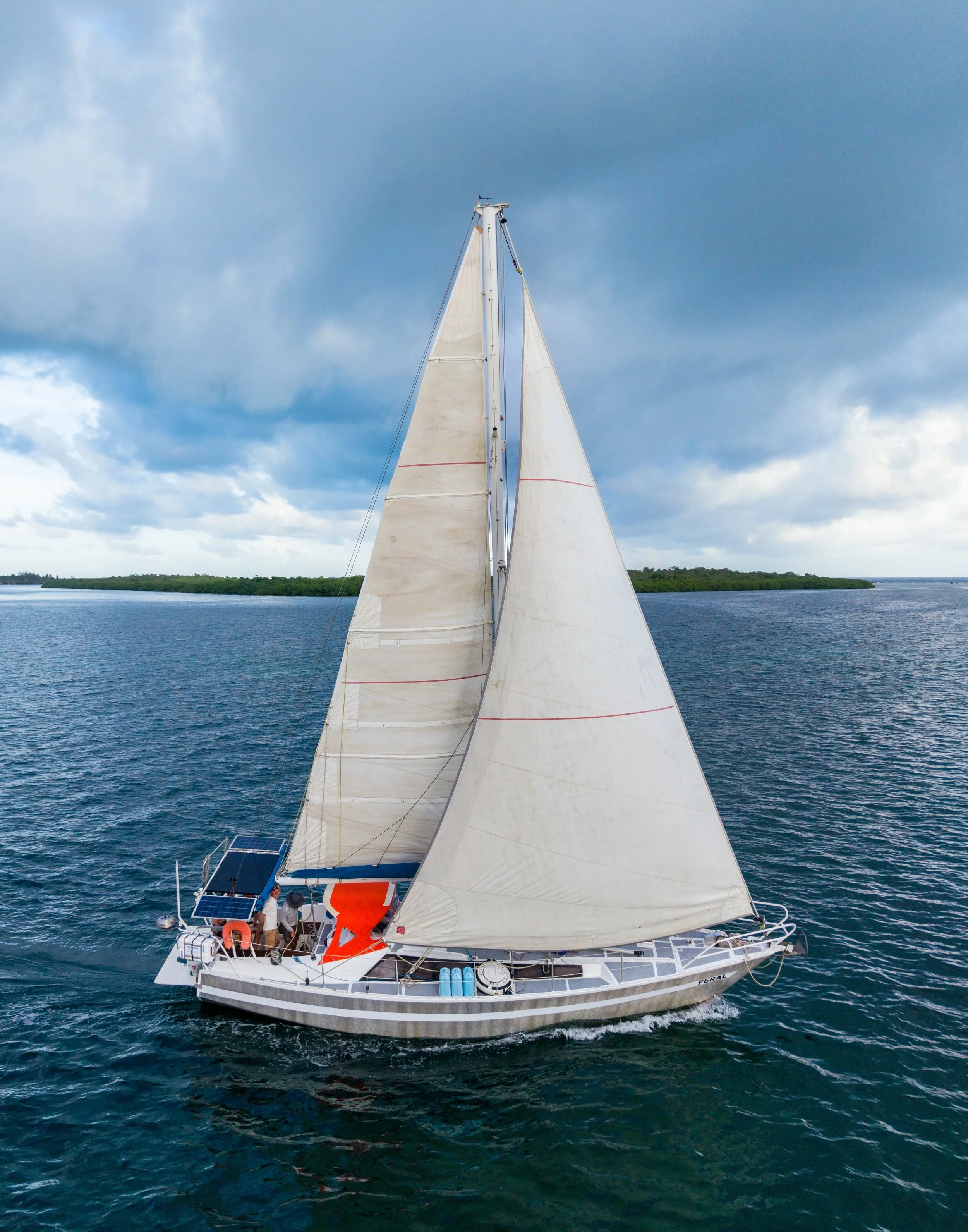 A white sailboat with two sails navigating on a body of water under a partly cloudy sky, with a landmass visible in the distance.