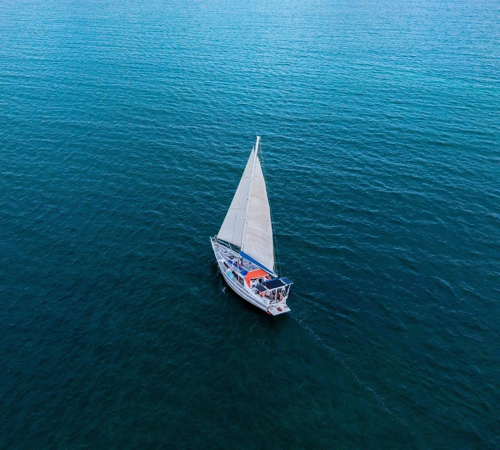 A sailboat with a white sail sailing on calm blue water from an aerial view.
