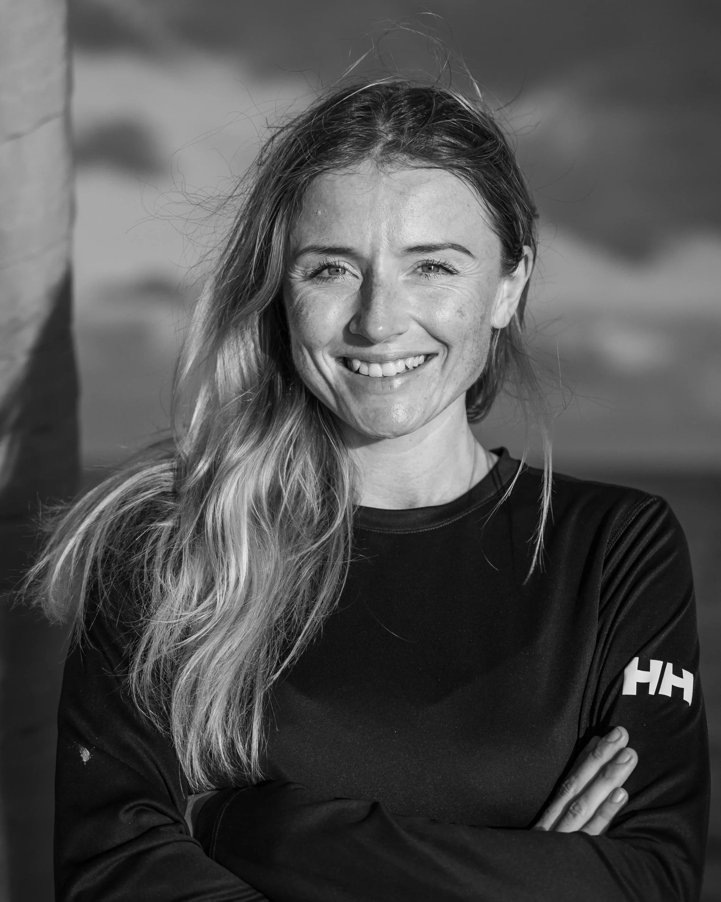 A young woman with long hair smiling outdoors, crossing her arms, wearing a black long-sleeve shirt with a logo on the sleeve.