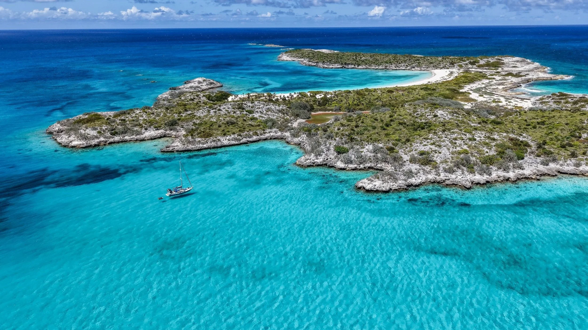 Aerial view of a tropical island with turquoise waters surrounding rocky and green land, a sailboat anchored near the shoreline, and a white sandy beach in the background.