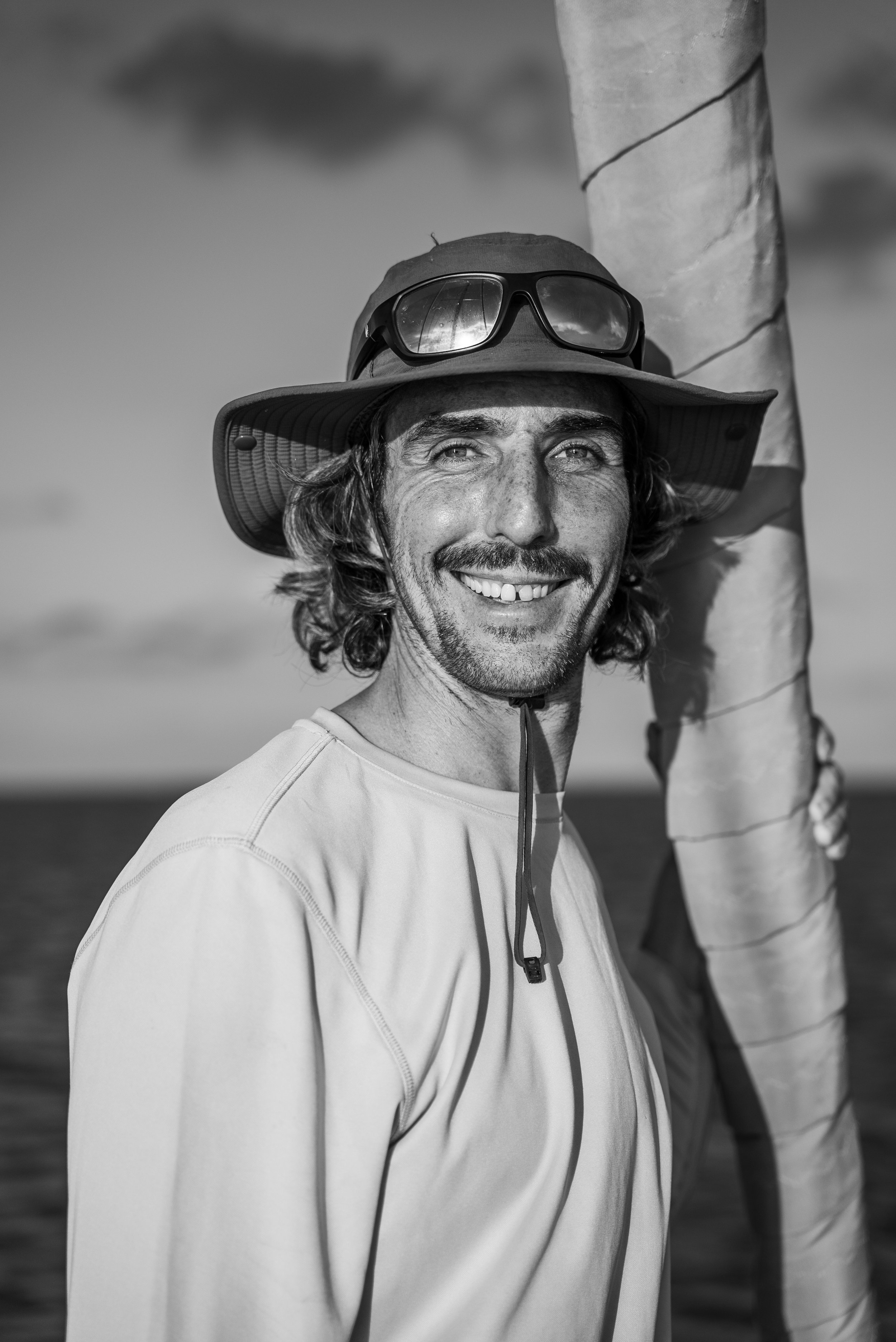 Black and white photo of a man smiling outdoors, wearing a wide-brimmed hat with sunglasses on top, and a light-colored long sleeve shirt.