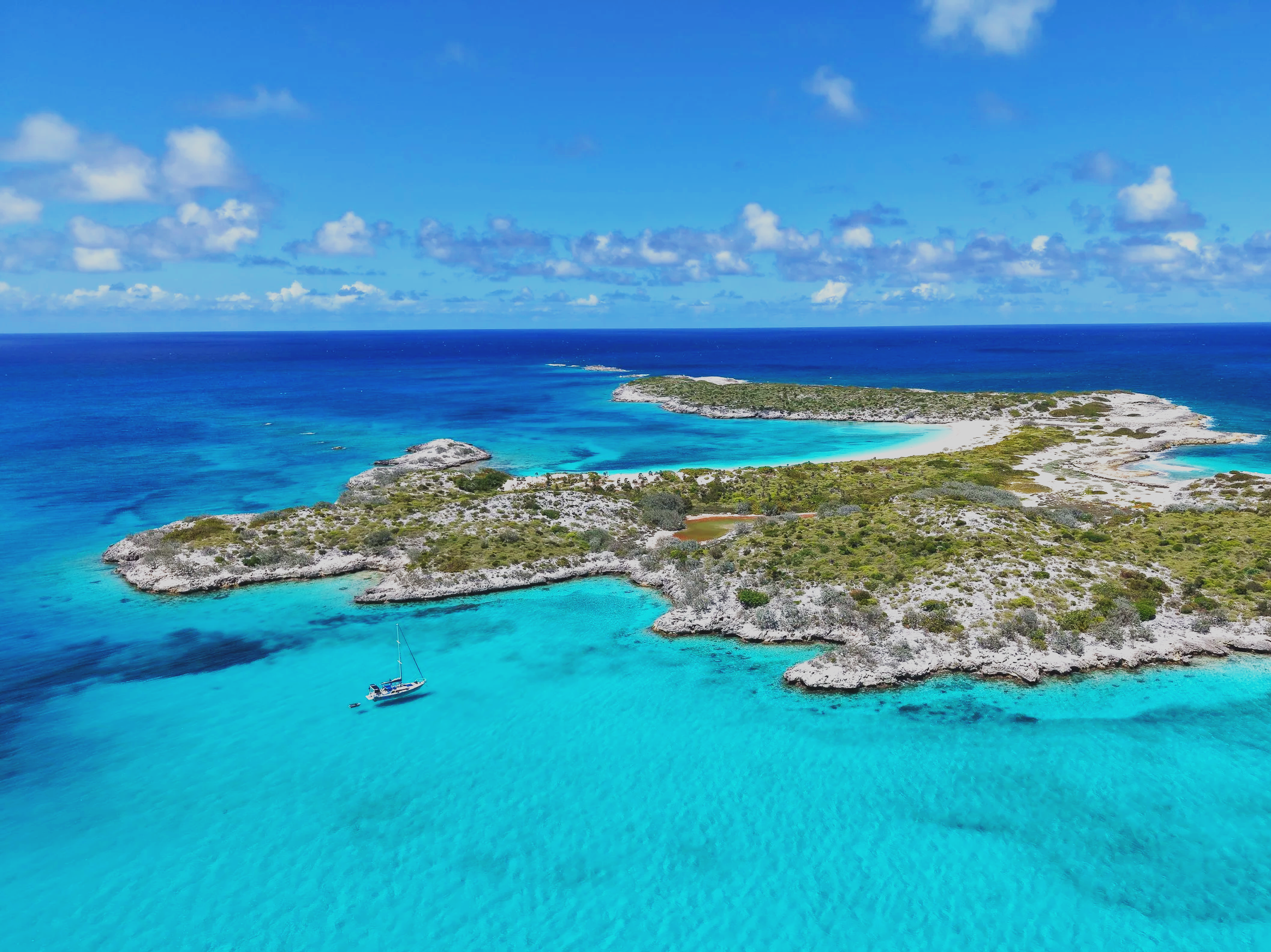 An aerial view of a tropical island with clear turquoise waters surrounding it, a sailboat anchored next to the island, and a sandy beach along the shoreline under a partly cloudy sky.