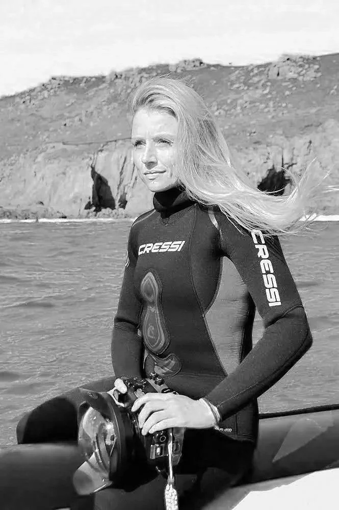 A woman in a wetsuit standing in a body of water with cliffs in the background.