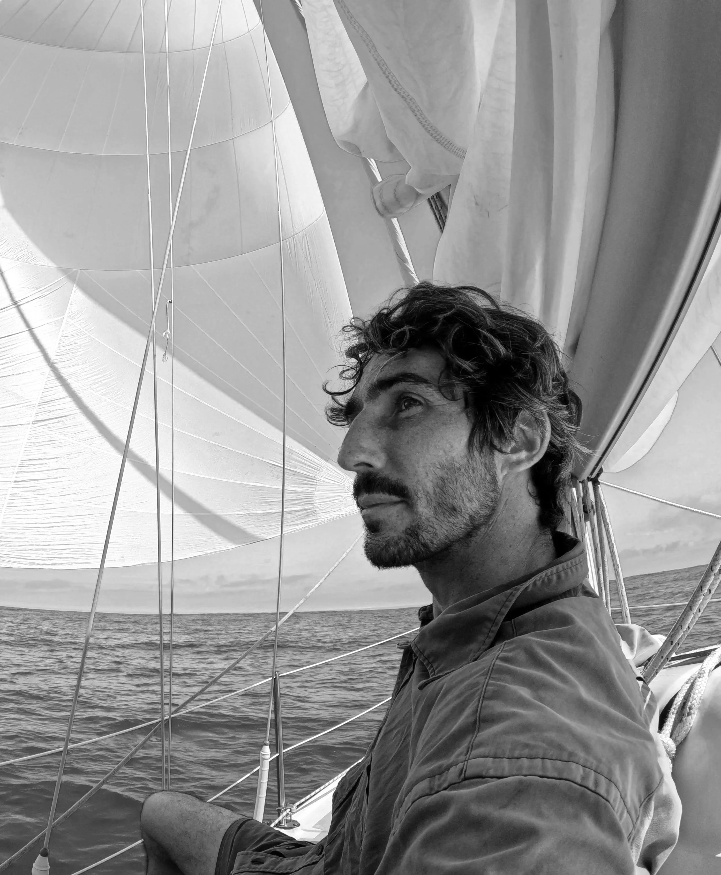 A man with curly hair and a beard sitting on a sailboat, looking to the right, with ocean water in the background and part of the boat's sail visible.