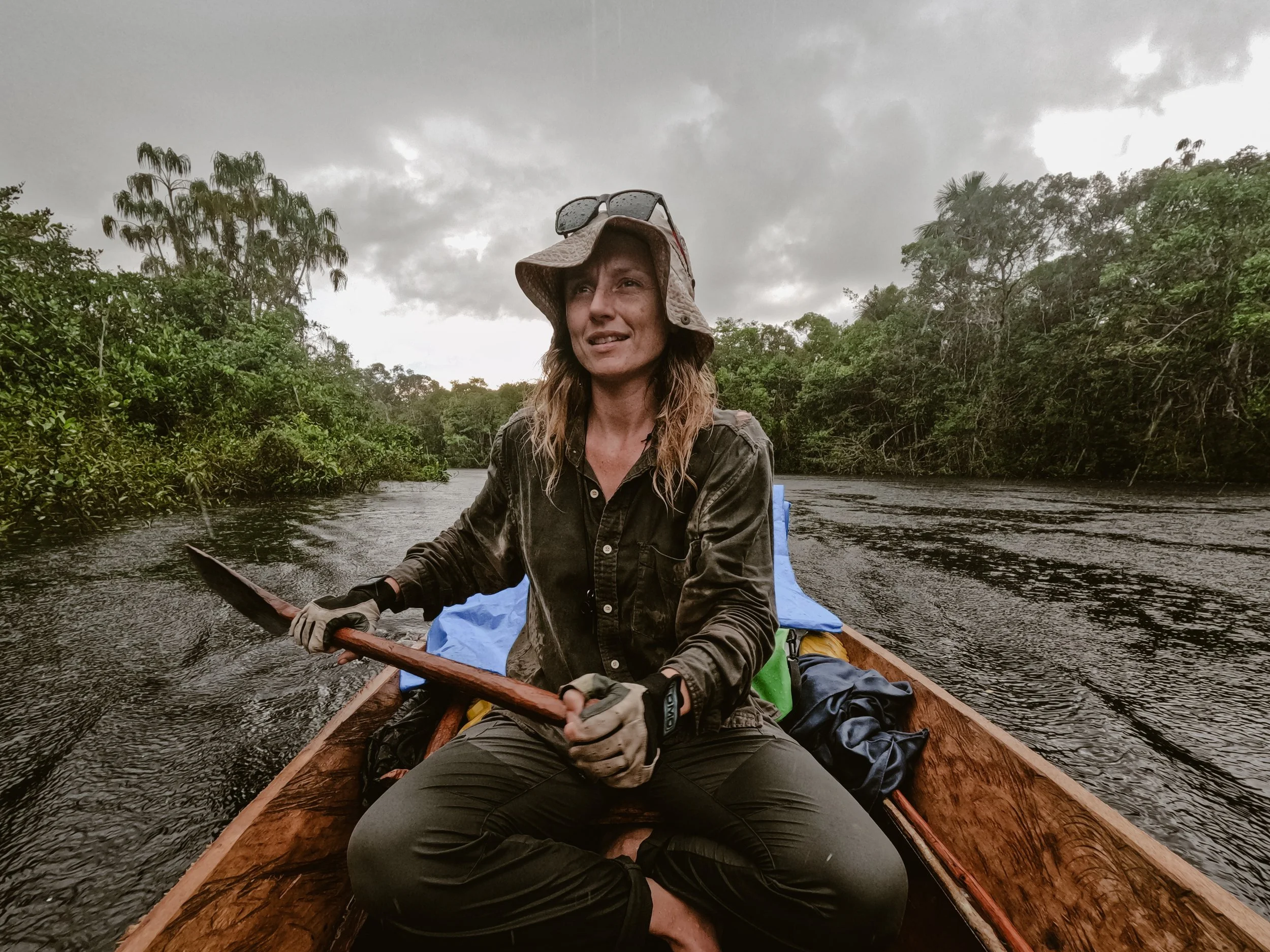A woman sitting in a wooden boat on a river, surrounded by lush green trees on cloudy weather, holding a paddle and wearing a wide-brimmed hat, sunglasses on her head, and outdoor clothing.