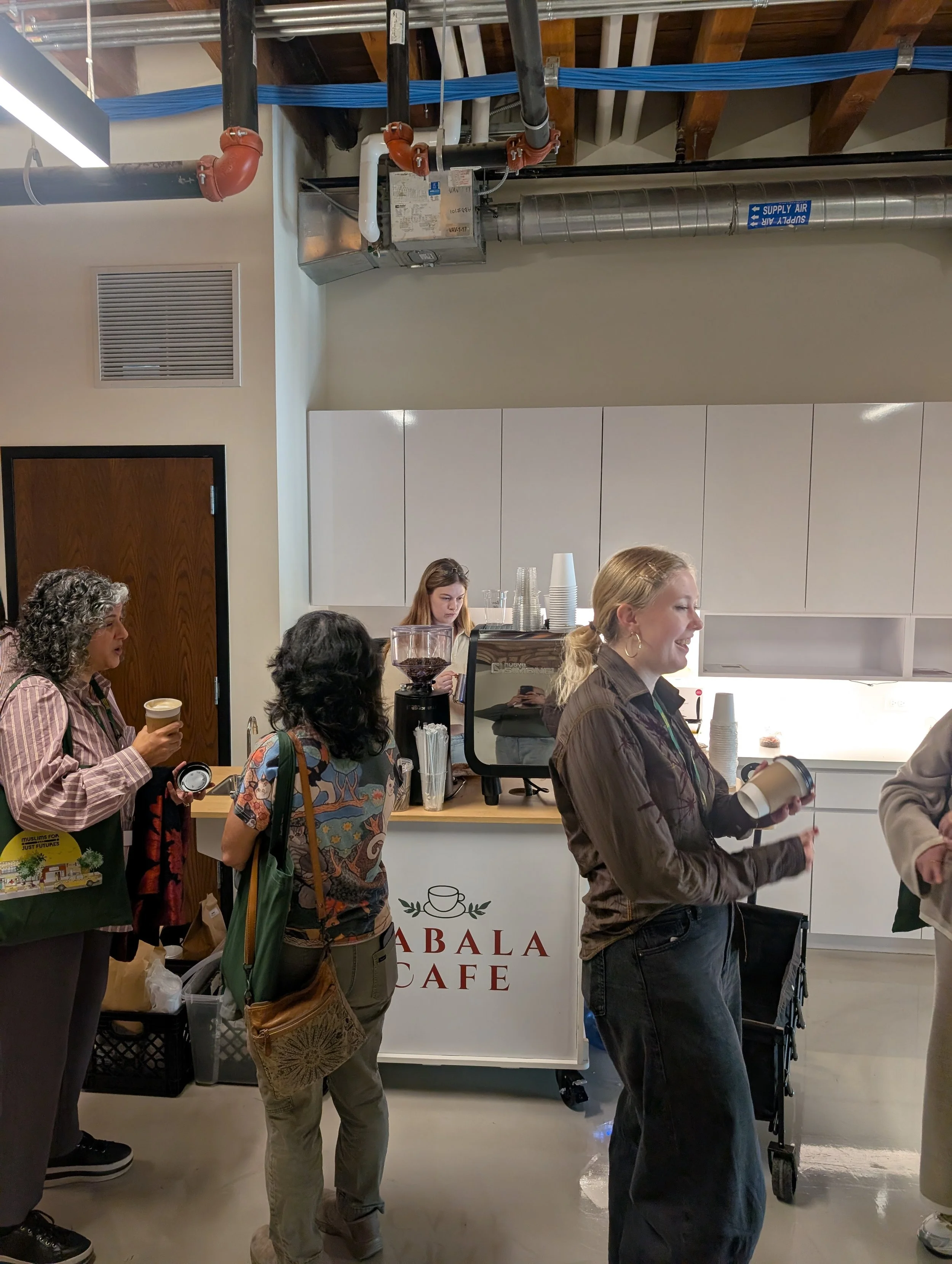 People gather at a coffee counter inside Ab Yala Cafe, with coffee equipment, cups, exposed HVAC pipes, and ductwork overhead.