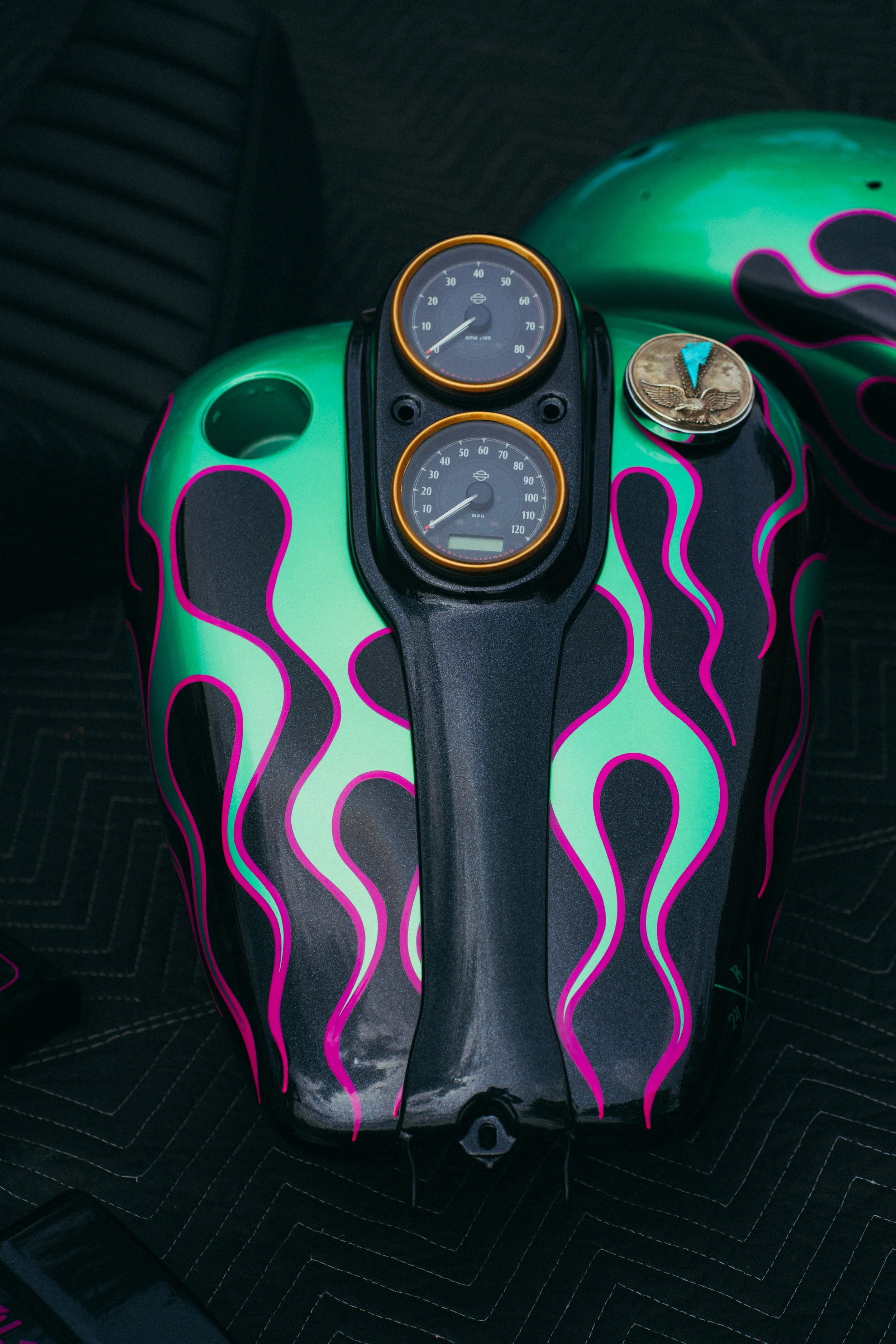 A motorcycle fuel tank painted with a black base, vibrant pink and green flame patterns, with two round gauges mounted in the center, a coin and a small pendant placed on the tank.