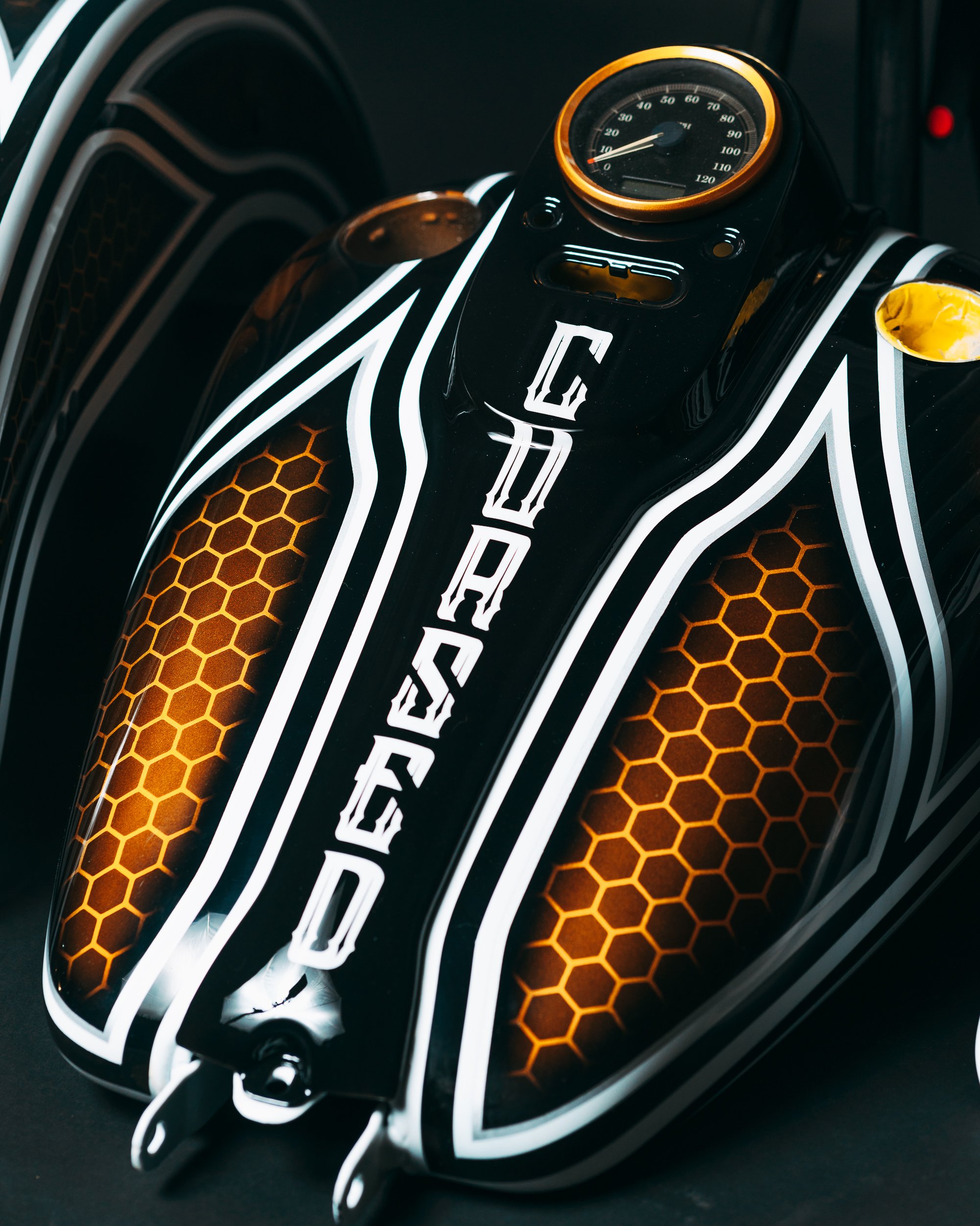 Close-up of a motorcycle gas tank with black, white, and orange honeycomb and stripe design, featuring the word 'GEARED' in stylized white letters and a round speedometer gauge on top.