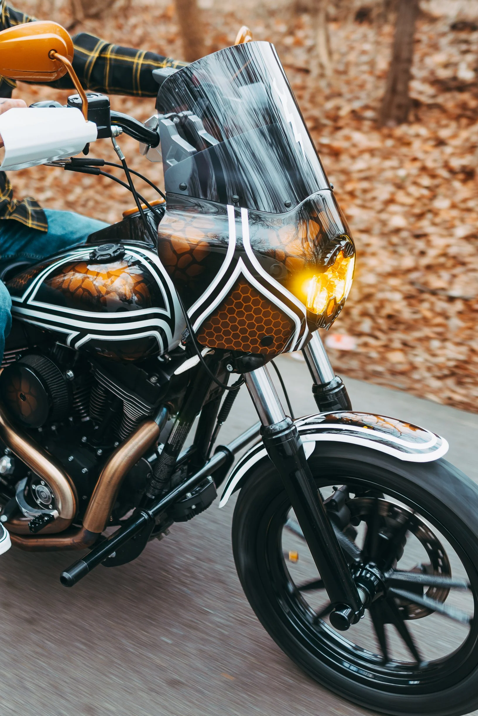 Close-up of a motorcycle with a black, white, and orange geometric design, riding on a trail with fallen leaves in the background.