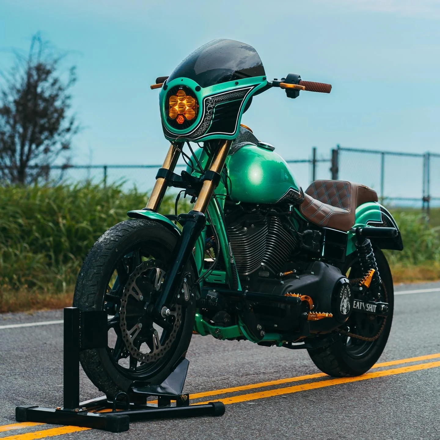 A custom green motorcycle with a brown leather seat parked on a road, with a black helmet on the handlebars, and a stand supporting the front wheel.