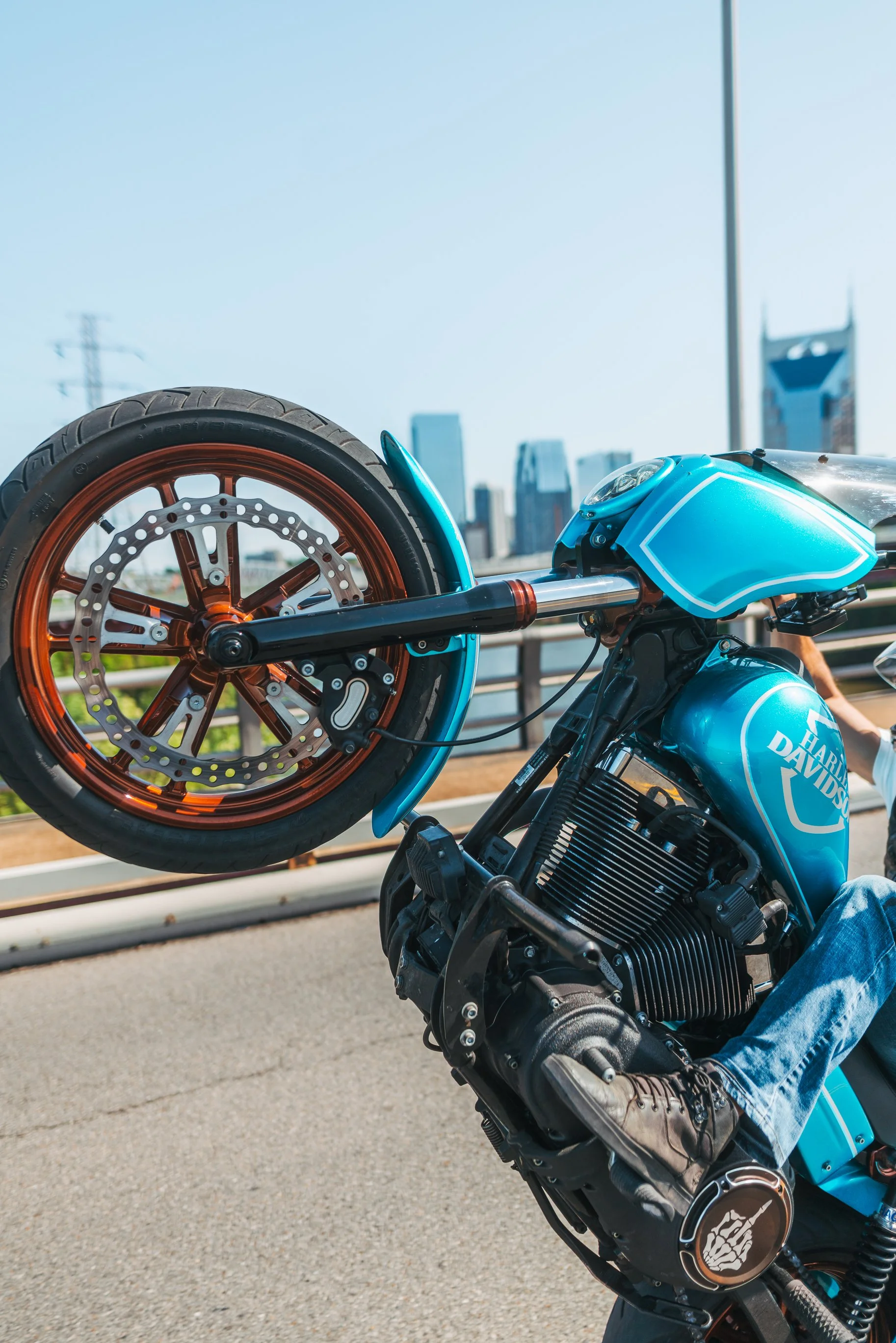 A person performing a wheelie on a blue Harley-Davidson motorcycle on a city street with a skyline in the background.