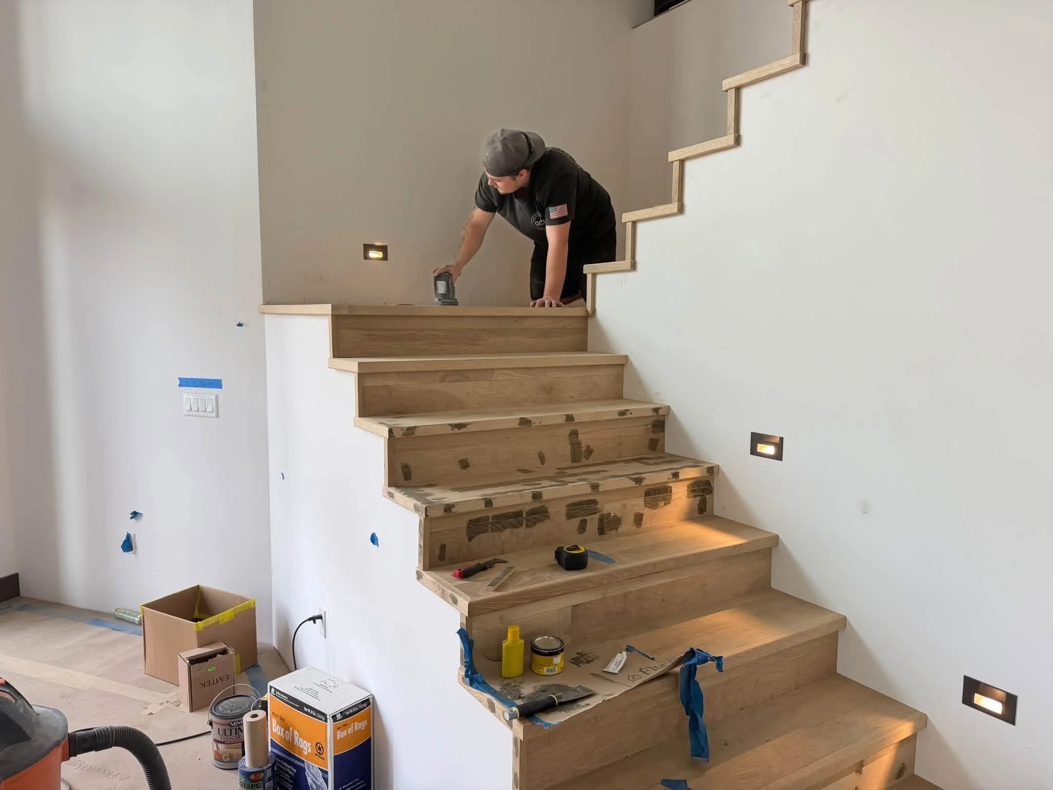 A person installing a wooden staircase in a home under construction, with various tools and supplies nearby.