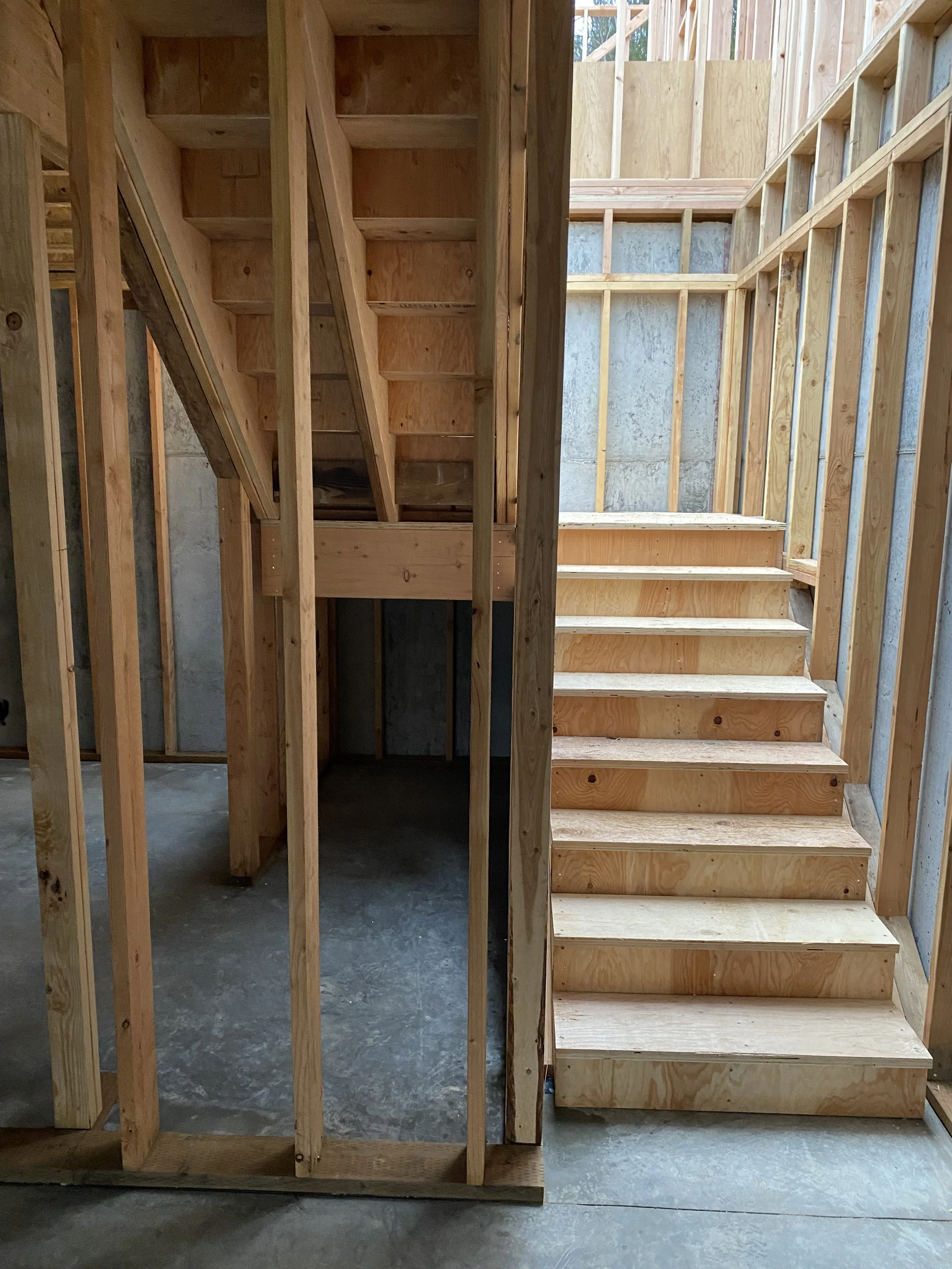 Interior view of a house under construction, showing wooden framing and staircase, with a concrete wall in the background.