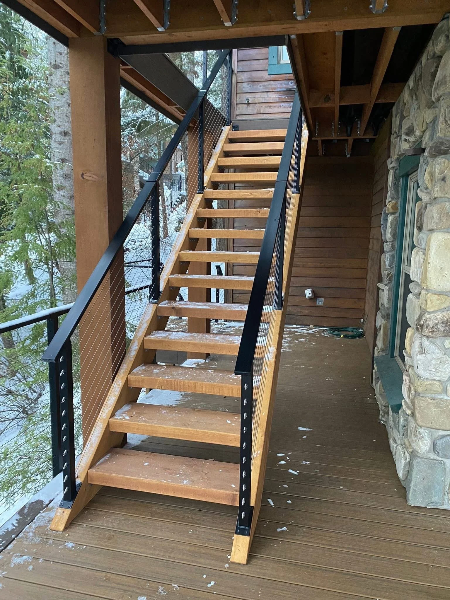 Unfinished wooden staircase with black metal railing on a wooden deck, leading to an upper level of a house with a stone and wood exterior, surrounded by trees.