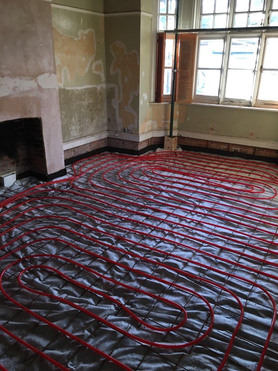 Underfloor heating pipes installation in a room with windows, unfinished walls, and a fireplace.