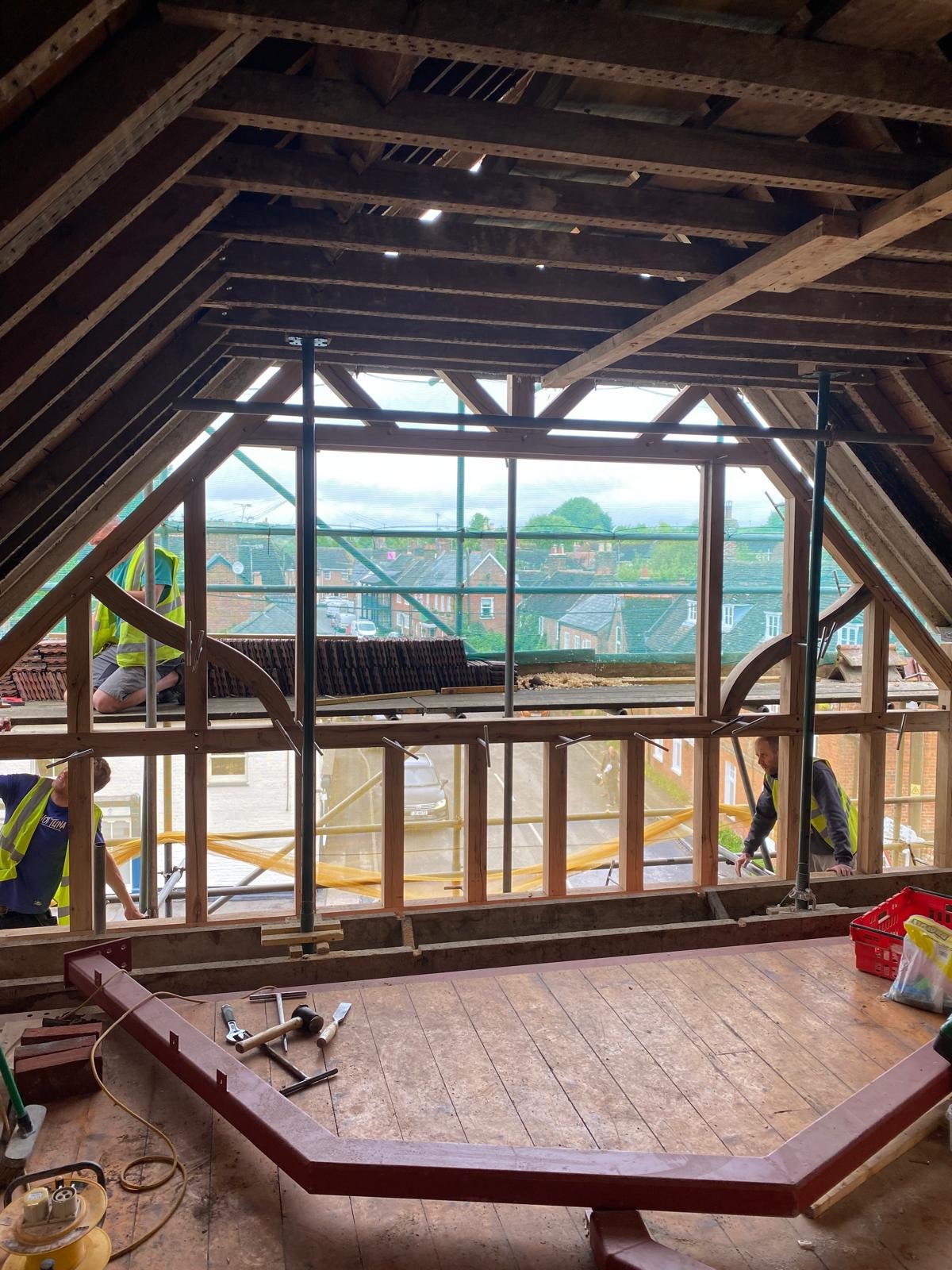 Interior view of an attic under construction with exposed wooden framing and scaffolding, showing construction workers installing windows and working on the roof.