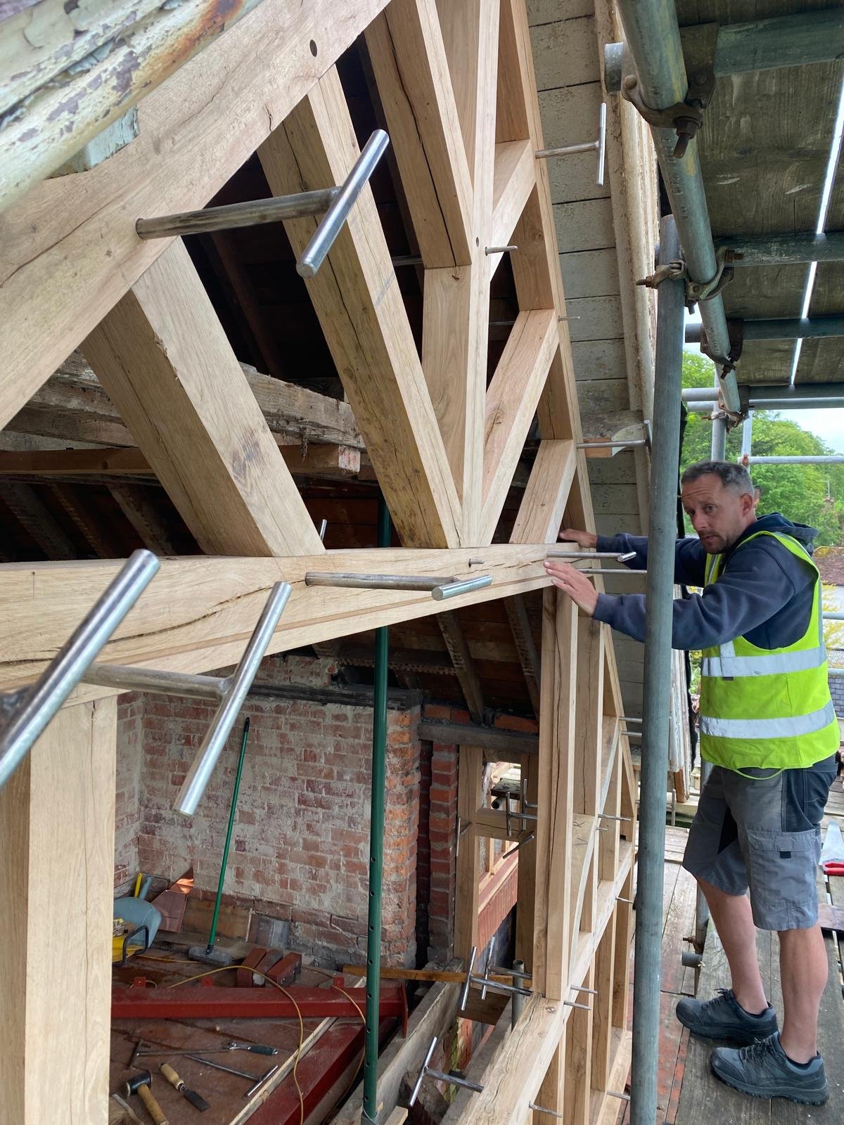 A worker in a safety vest and shorts is working on a wooden frame for a building under construction, with metal scaffolding and brick wall visible.