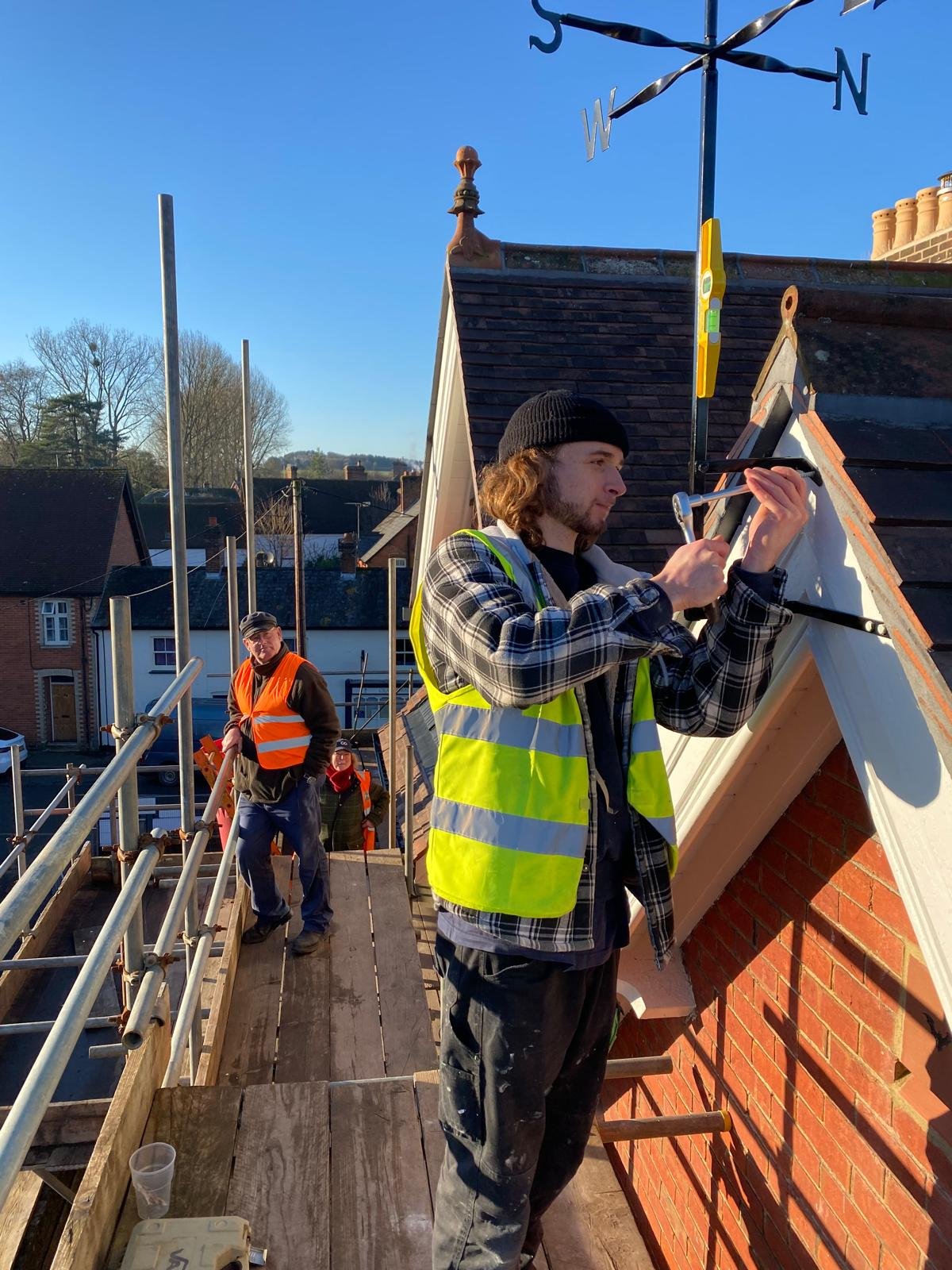 Construction workers installing a weather vane on the roof of a building. One worker in a yellow safety vest is using a tool on the weather vane, while two other workers in orange safety vests are seen further back on the scaffold. The sky is clear a
