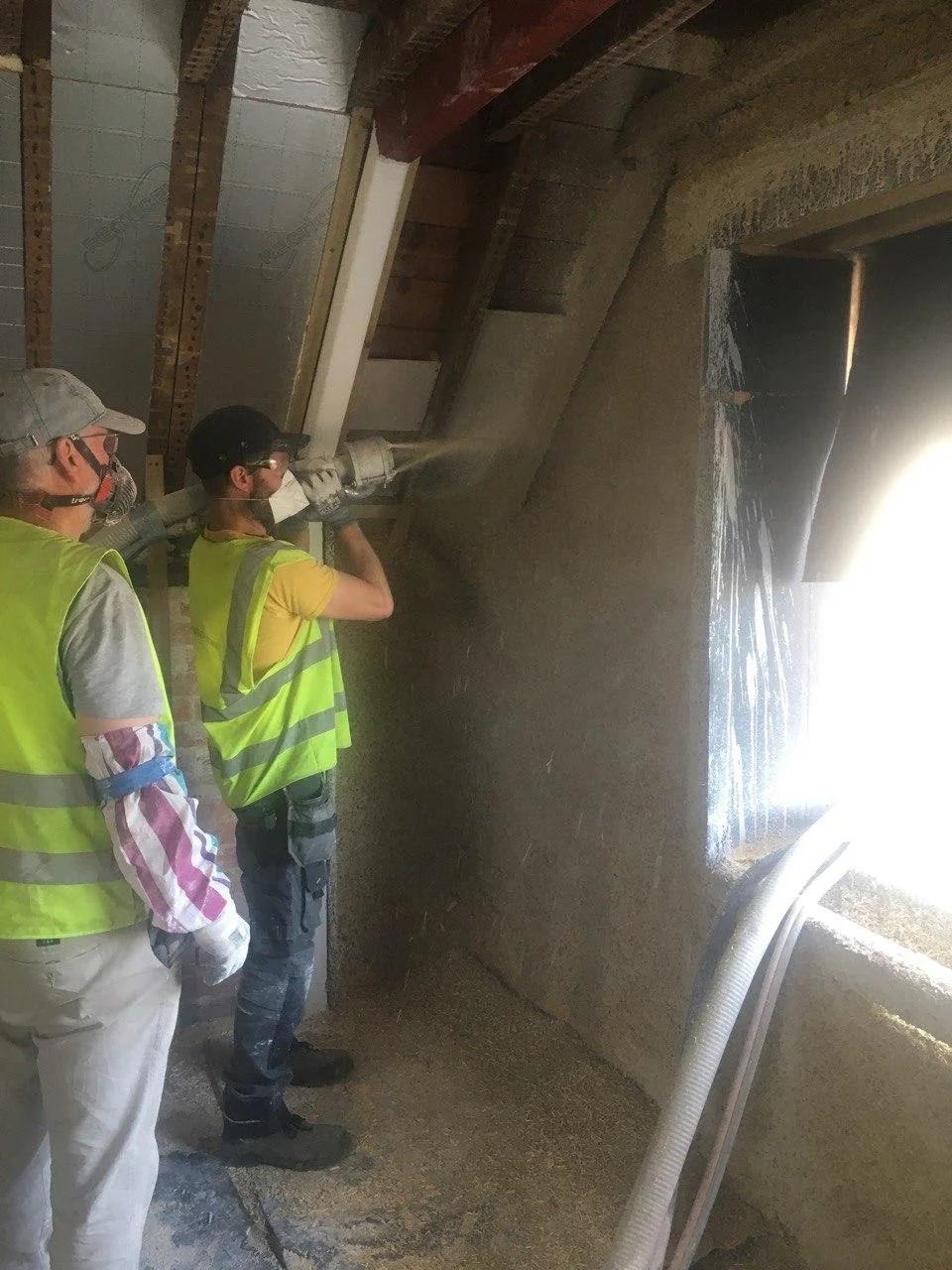 Two construction workers in yellow safety vests and masks pouring concrete inside a building.