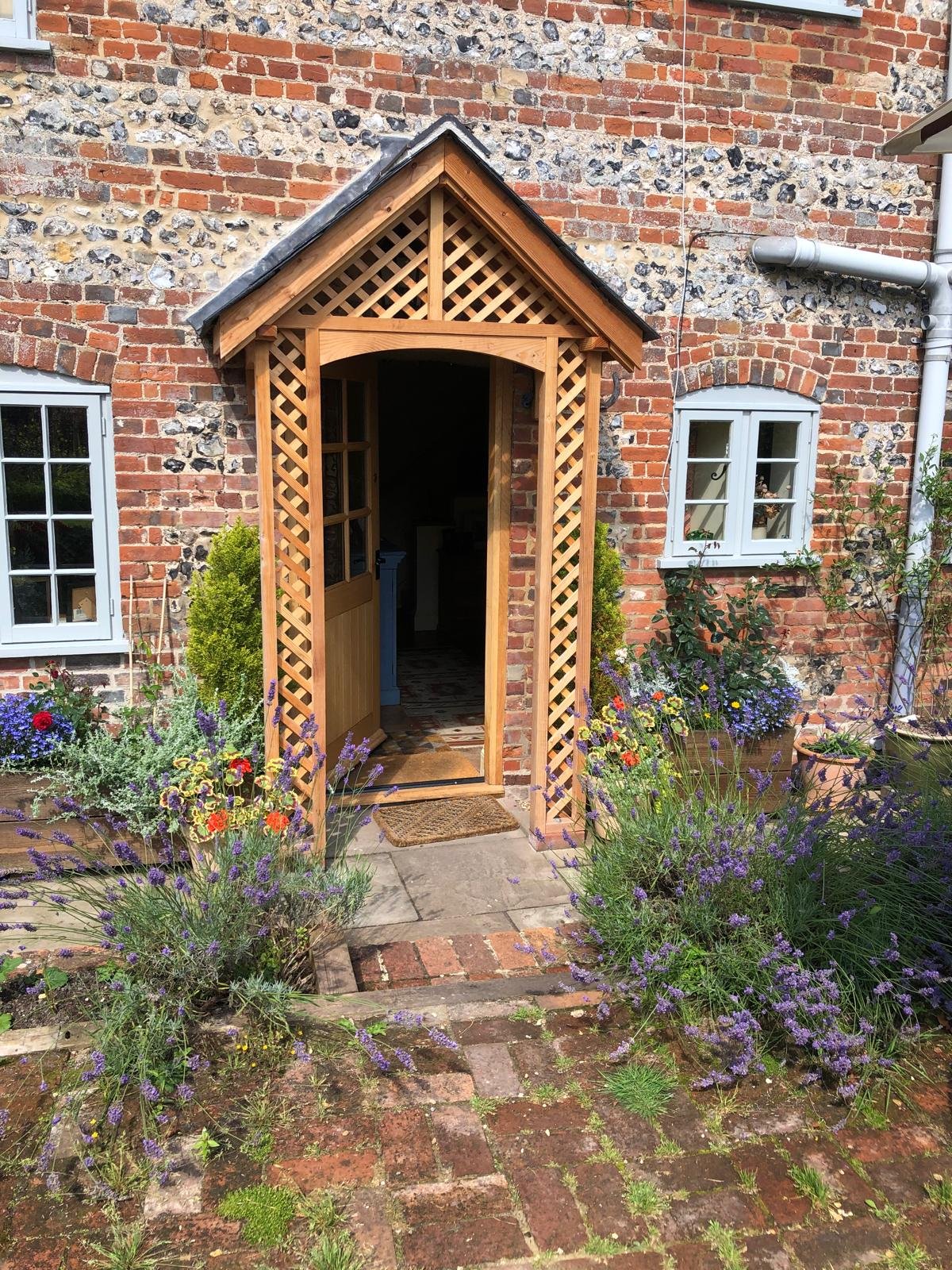 Front view of a brick house entryway with a wooden lattice-covered porch, surrounded by colorful flowers and plants.