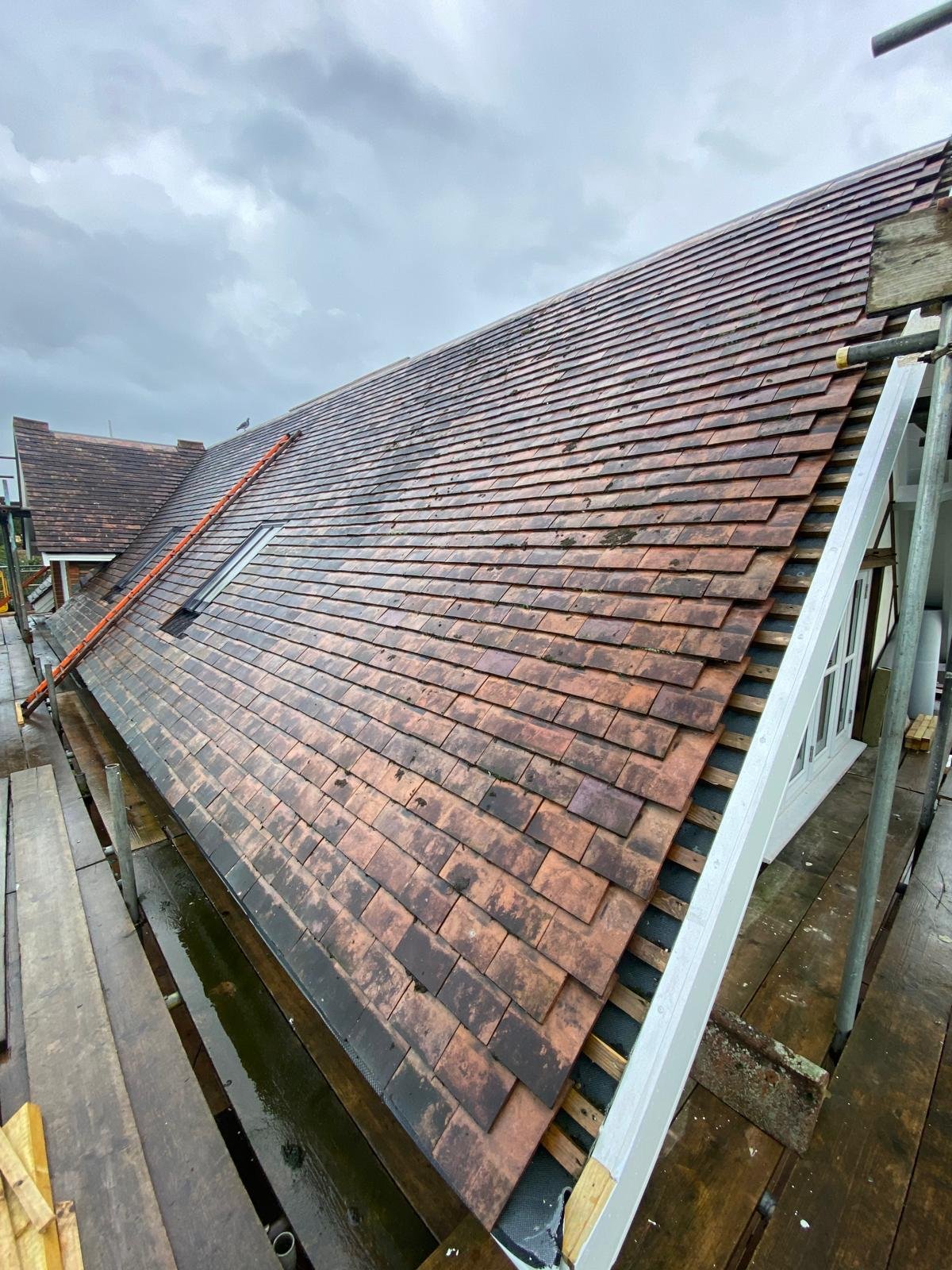 A steep residential roof under construction with red-brown tiles and a skylight, on a cloudy day.