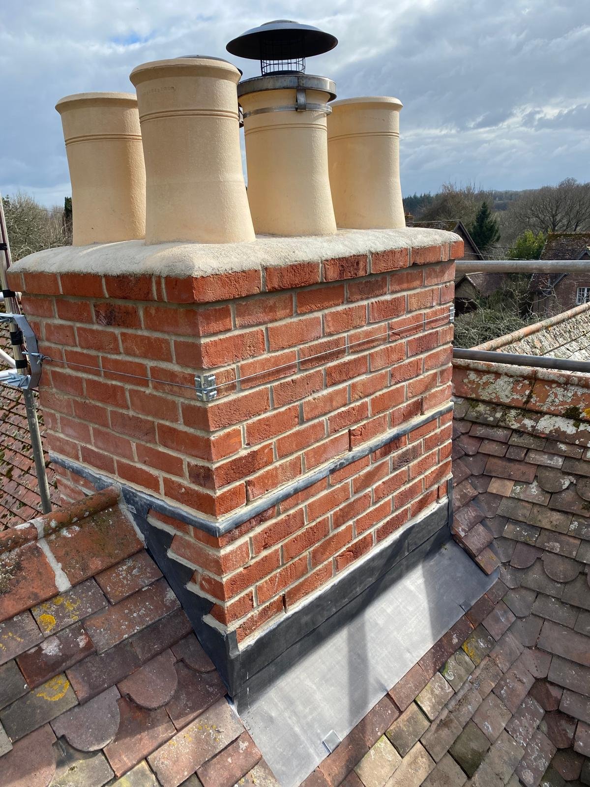 Close-up view of a brick chimney on a rooftop, with four chimney pots and a rain cap, surrounded by roofing tiles and scaffolding, with trees and houses in the background.