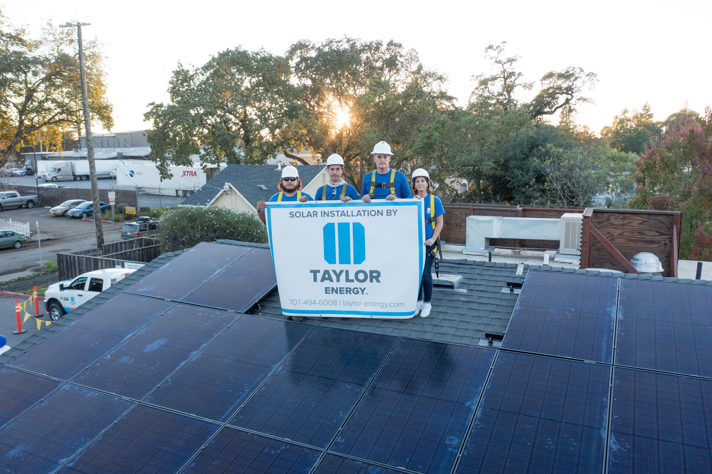 Four workers standing on a rooftop with solar panels, holding a sign for Taylor Energy, with trees and an industrial area in the background.