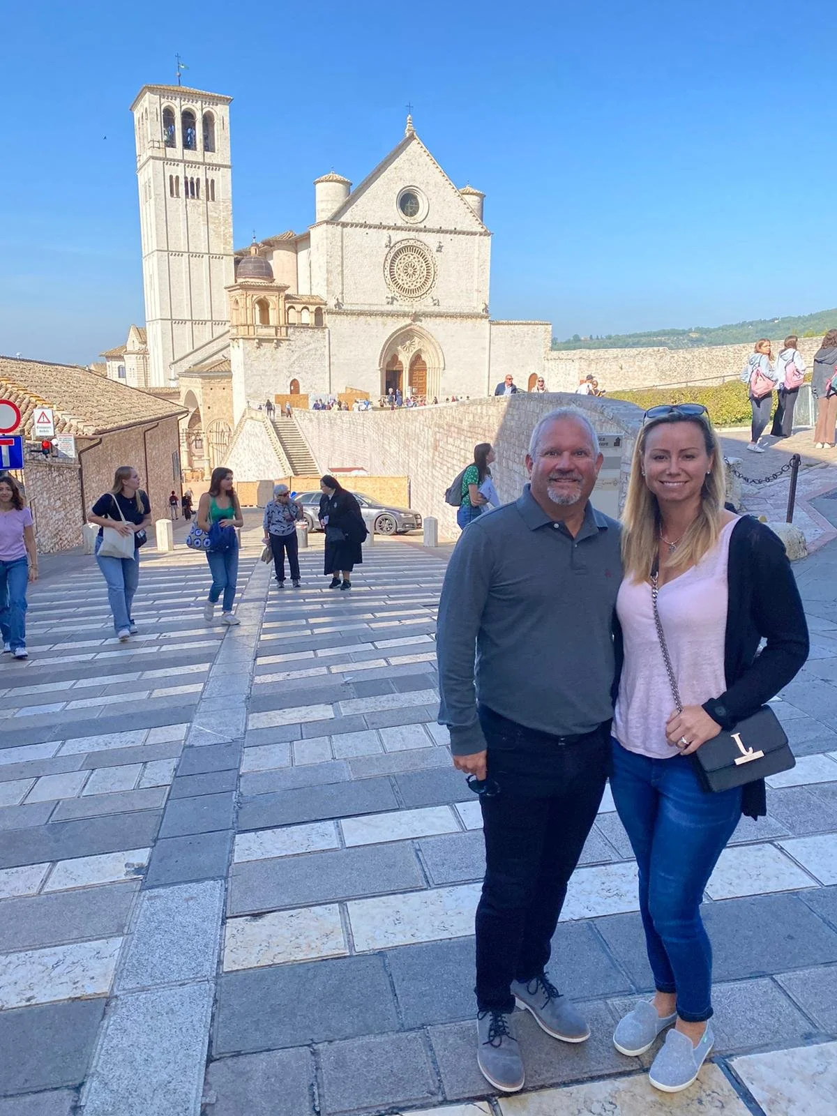 A man and woman standing together in front of a historic stone church with a bell tower, during daytime with a blue sky. Several people are walking and gathering around them.