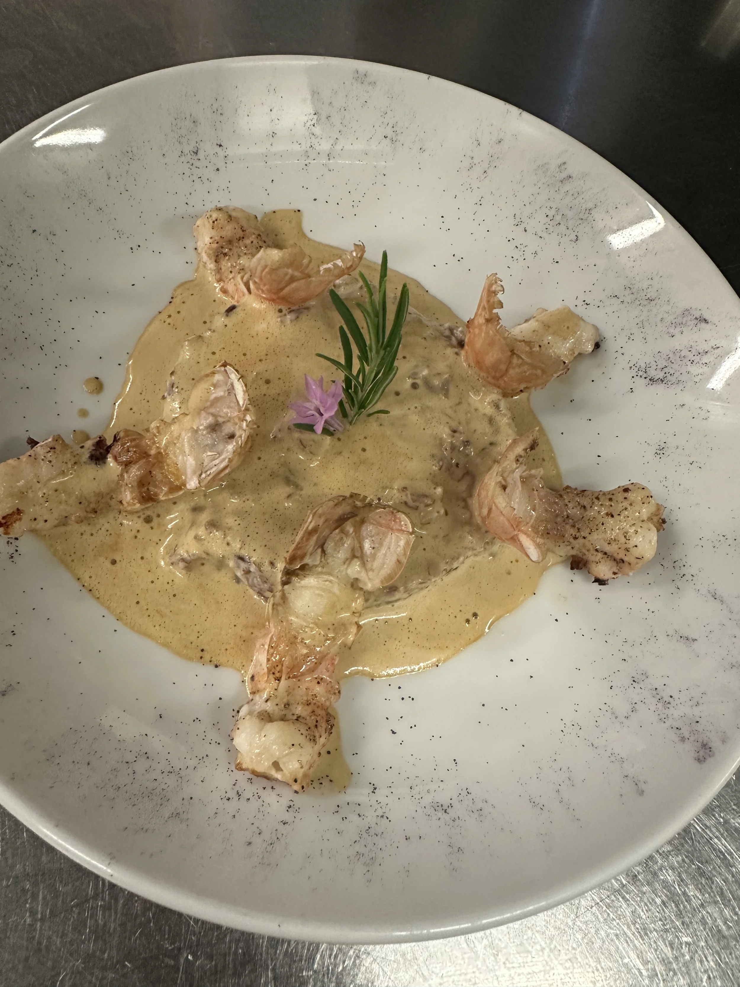 Seafood dish with shrimp in a creamy sauce, garnished with a sprig of rosemary and a pink edible flower on a white plate